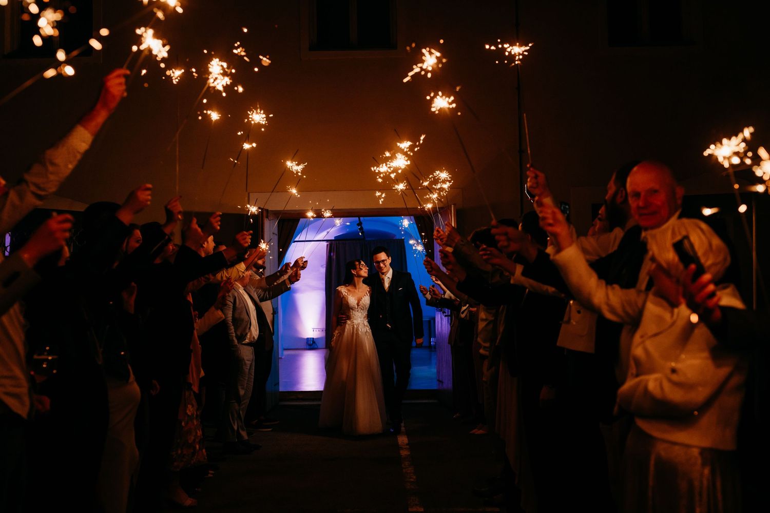 Newlyweds exit through sparkler tunnel at night wedding celebration with guests holding sparklers.