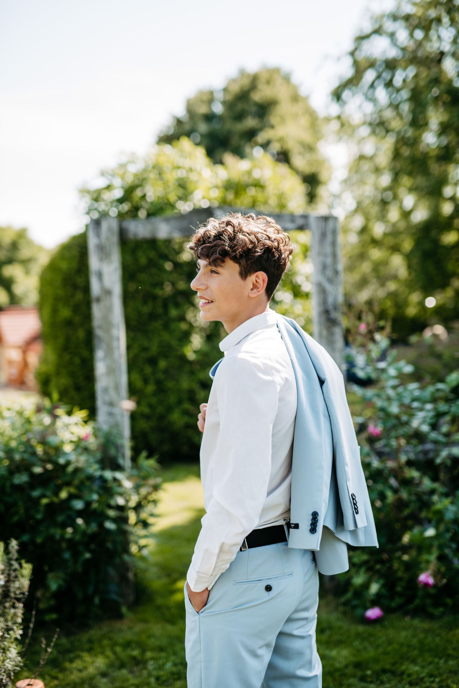 Side profile fashion pose in a garden setting wearing crisp white shirt and light blue suit.