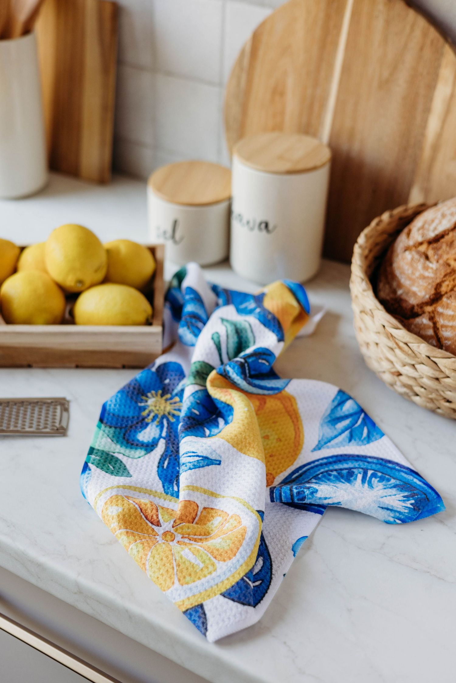 Colorful lemon-print kitchen towel on marble countertop with fresh lemons, bread basket, and wooden accessories.