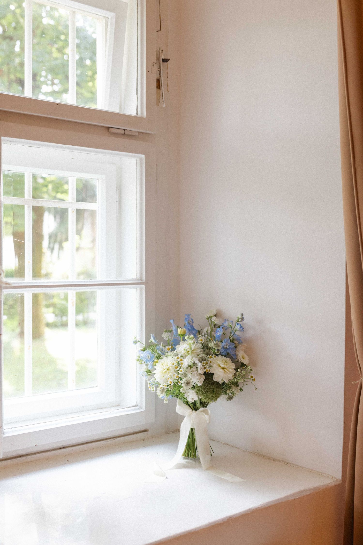 A delicate bridal bouquet of white and blue flowers tied with ribbon sits on a bright windowsill in a softly lit room.