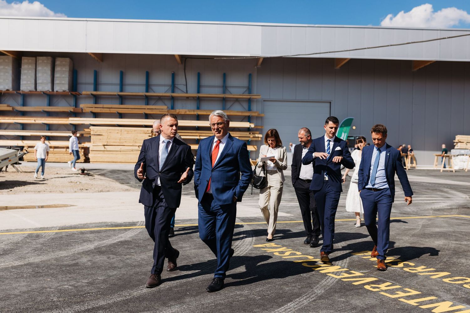 Business executives in suits walking confidently outside a modern industrial warehouse facility on a sunny day.