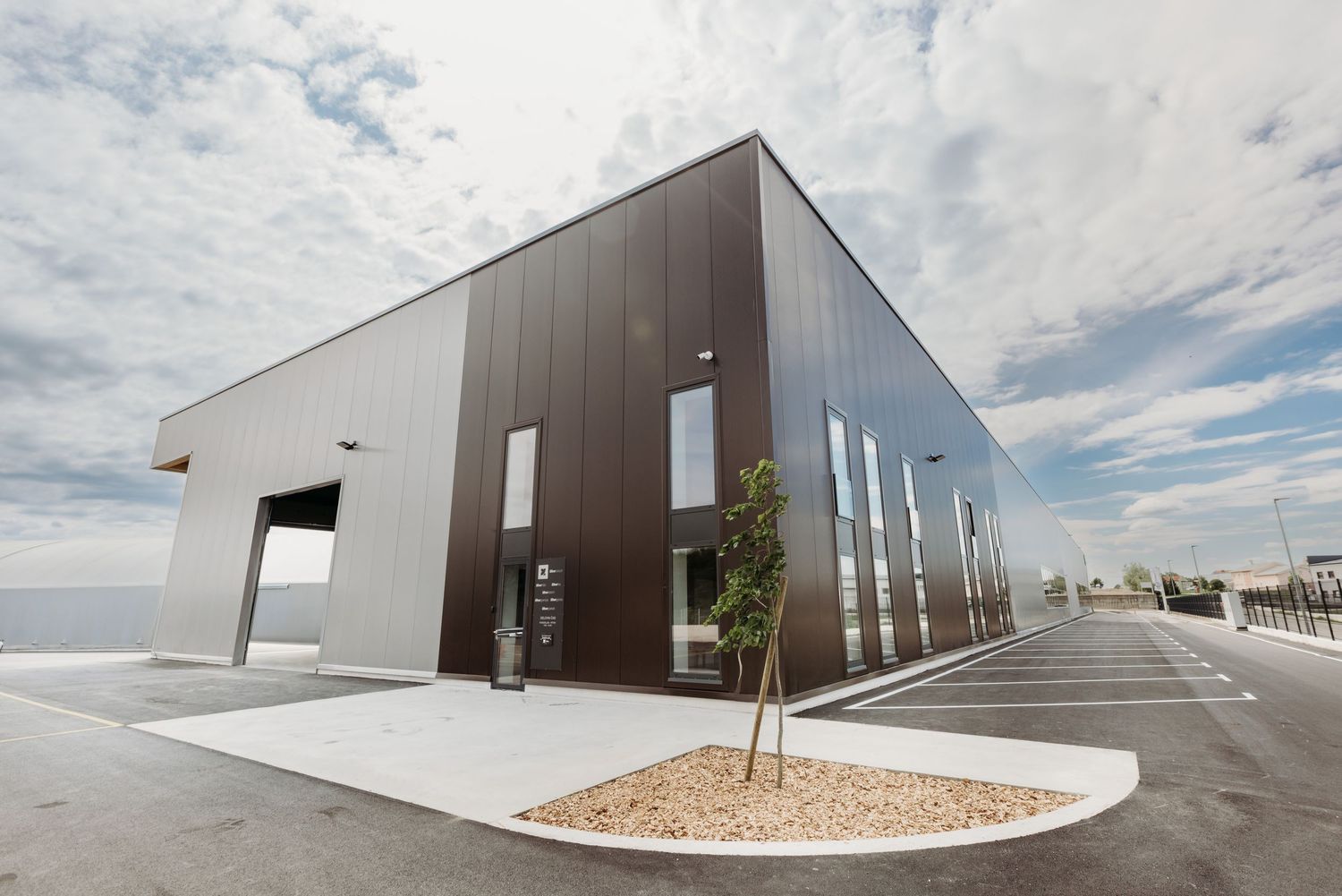Modern industrial warehouse building with dark brown metal cladding, large windows, and a young tree in the foreground.