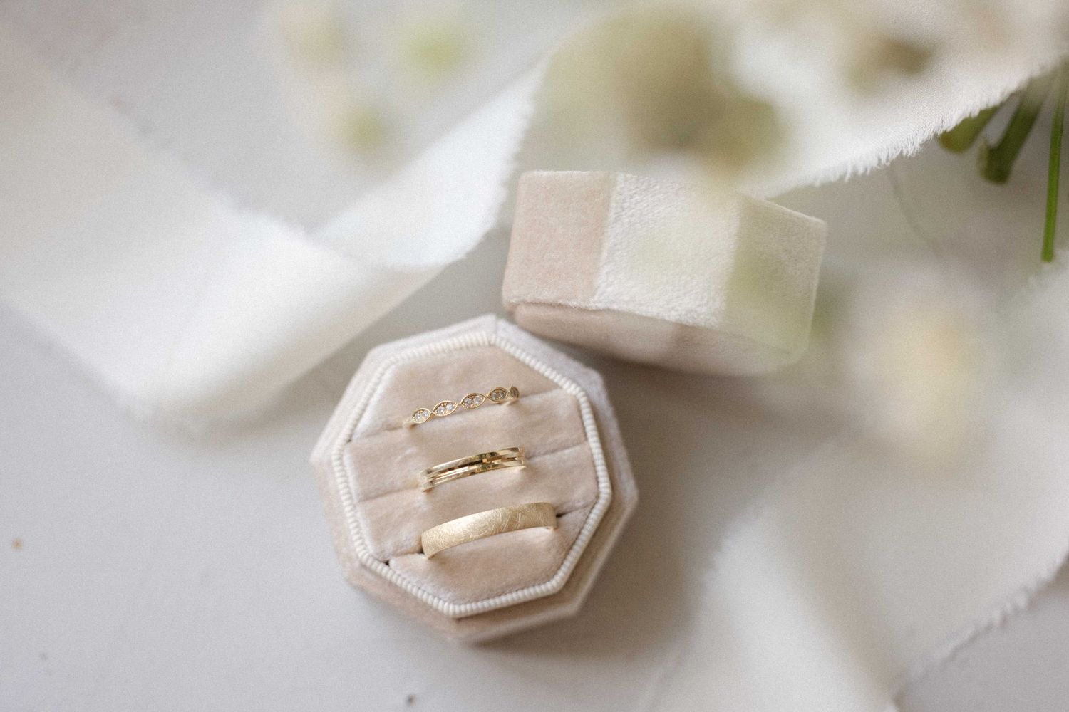 Two gold wedding bands displayed in an octagonal velvet ring box surrounded by white ribbon and flowers.