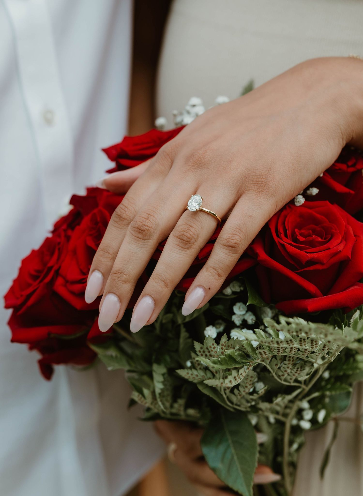 A hand wearing an engagement ring rests on a bouquet of red roses with delicate baby's breath accents.
