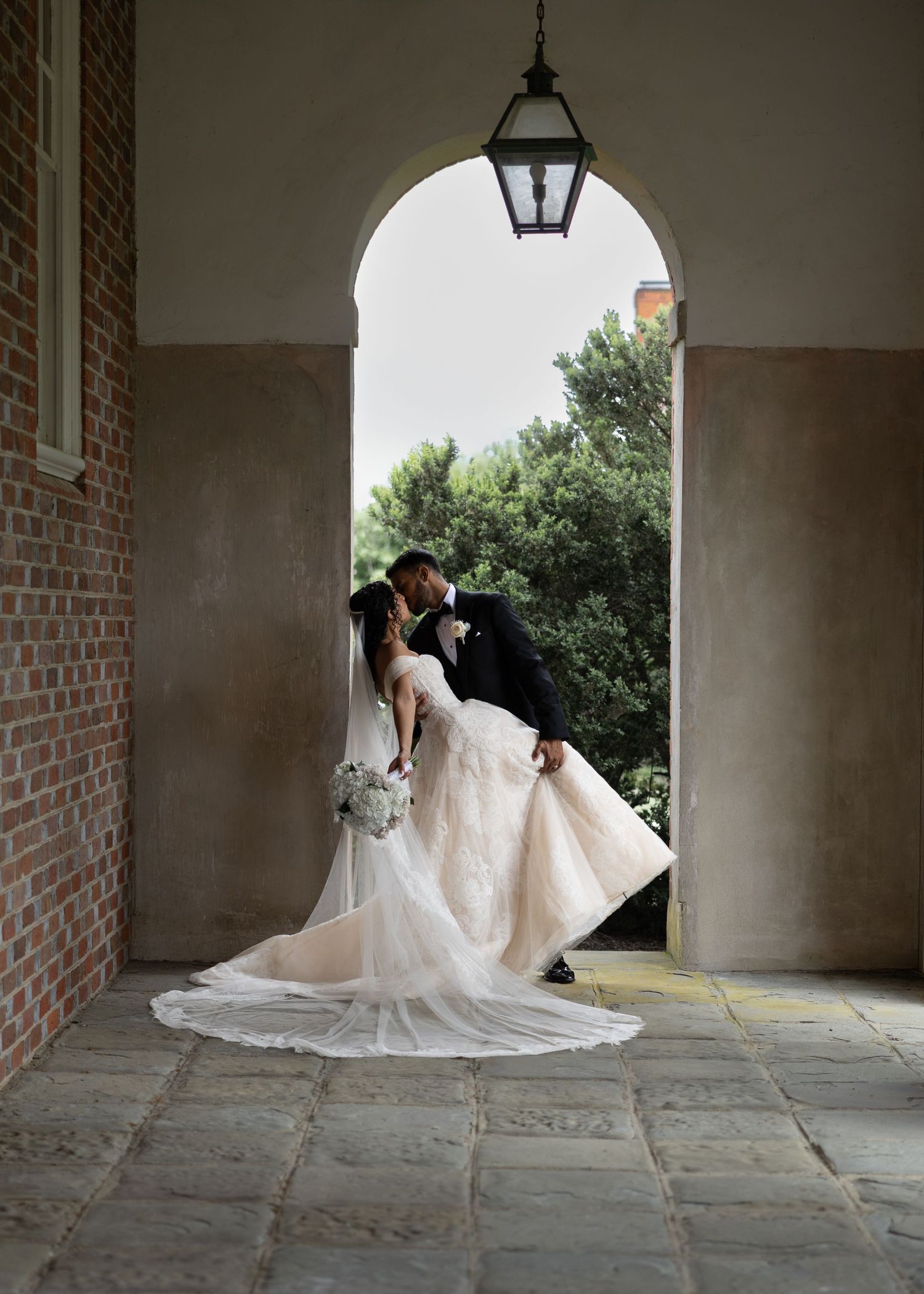 Bride and groom share a romantic dip kiss framed by an elegant arched doorway with a vintage lantern overhead.