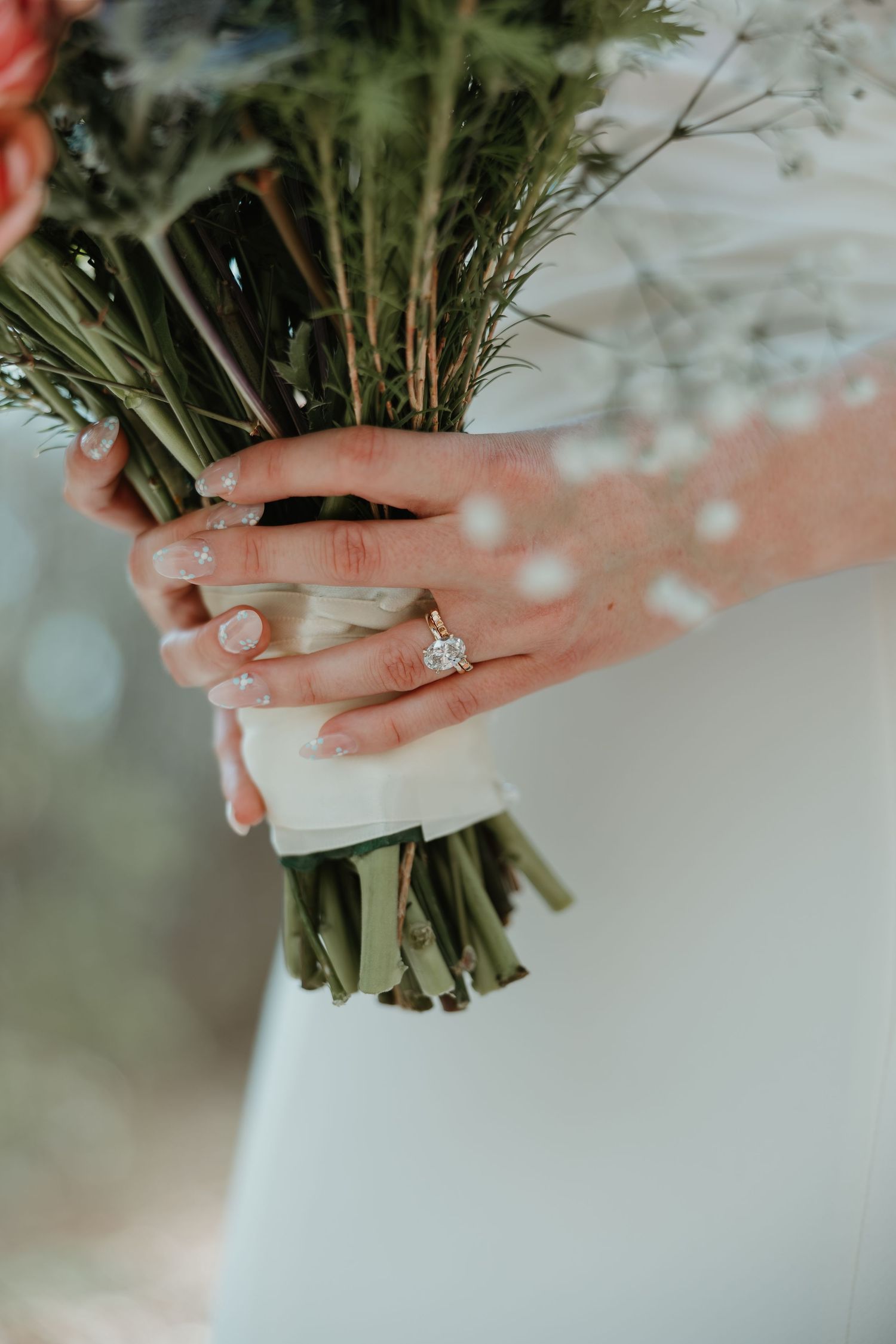 Bride holding a green floral bouquet wrapped in white ribbon, wearing a solitaire diamond engagement ring on her finger.