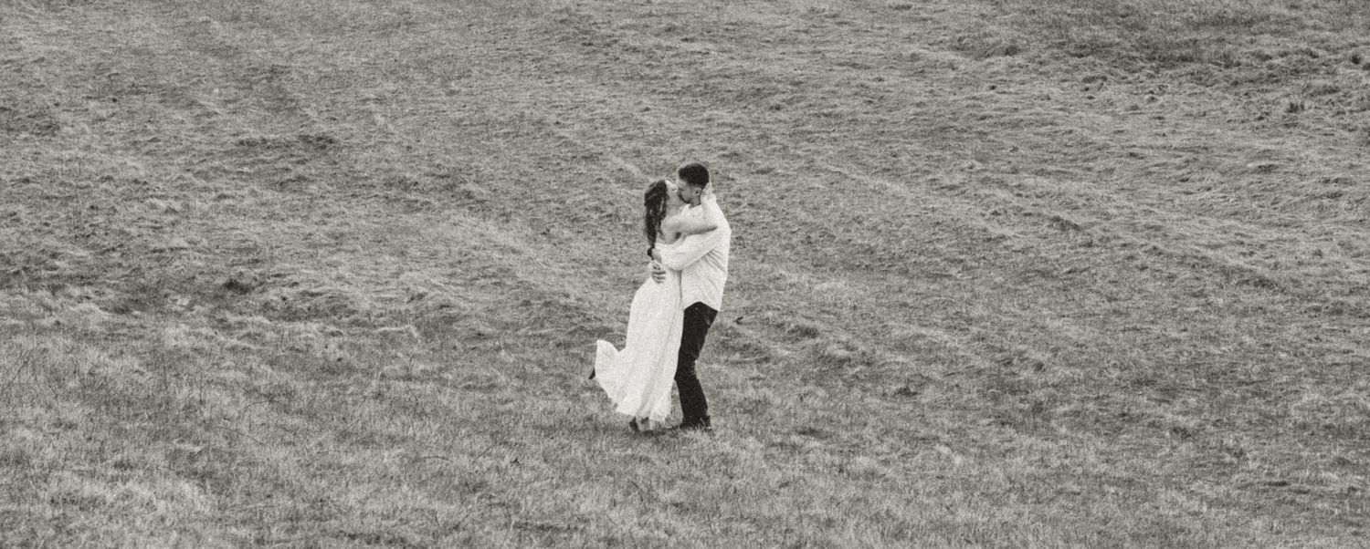 Black and white photo of a couple embracing in a vast grassy field, bride in white gown and groom in dark suit.