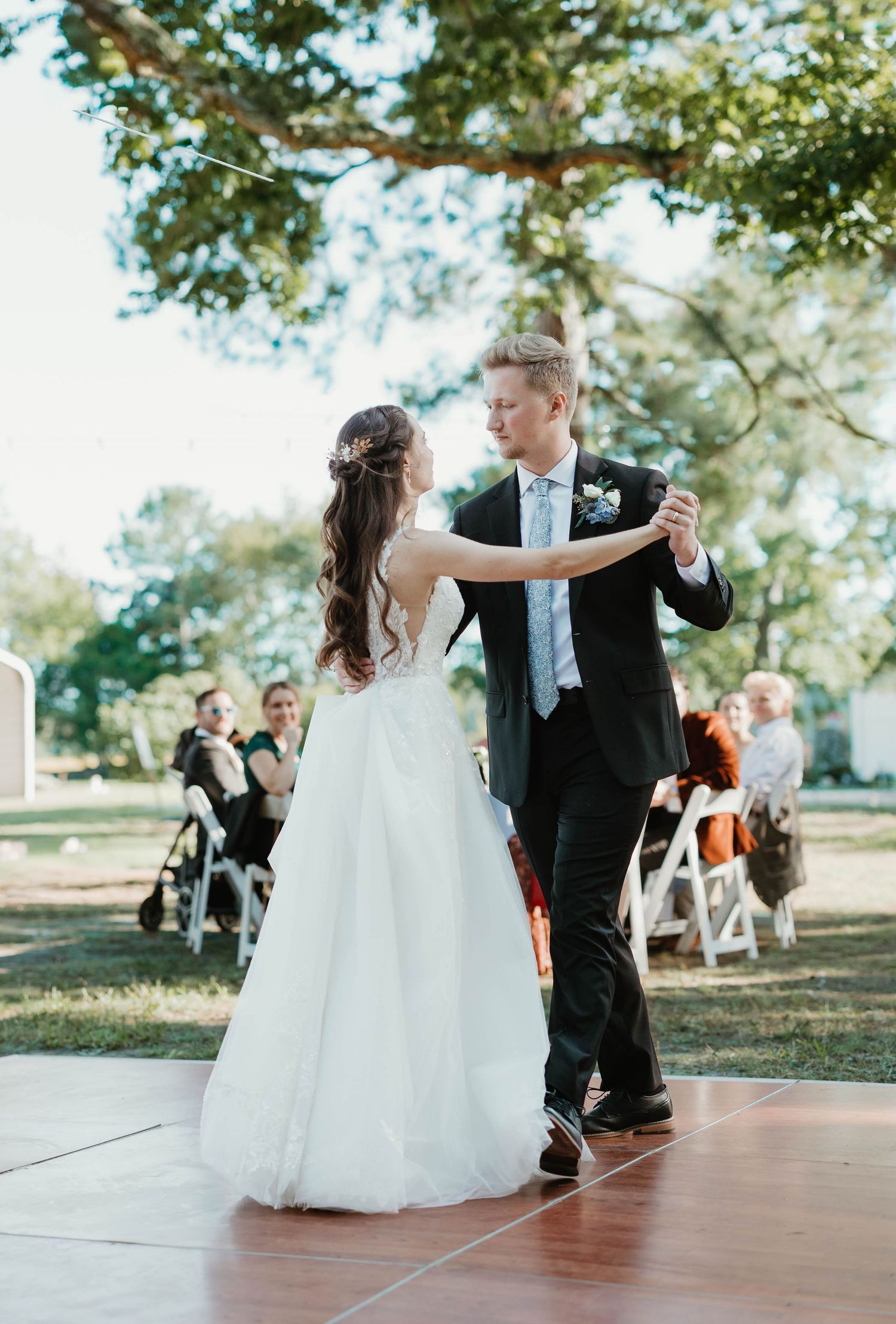 A couple shares their first dance as newlyweds on an outdoor dance floor surrounded by trees and guests.
