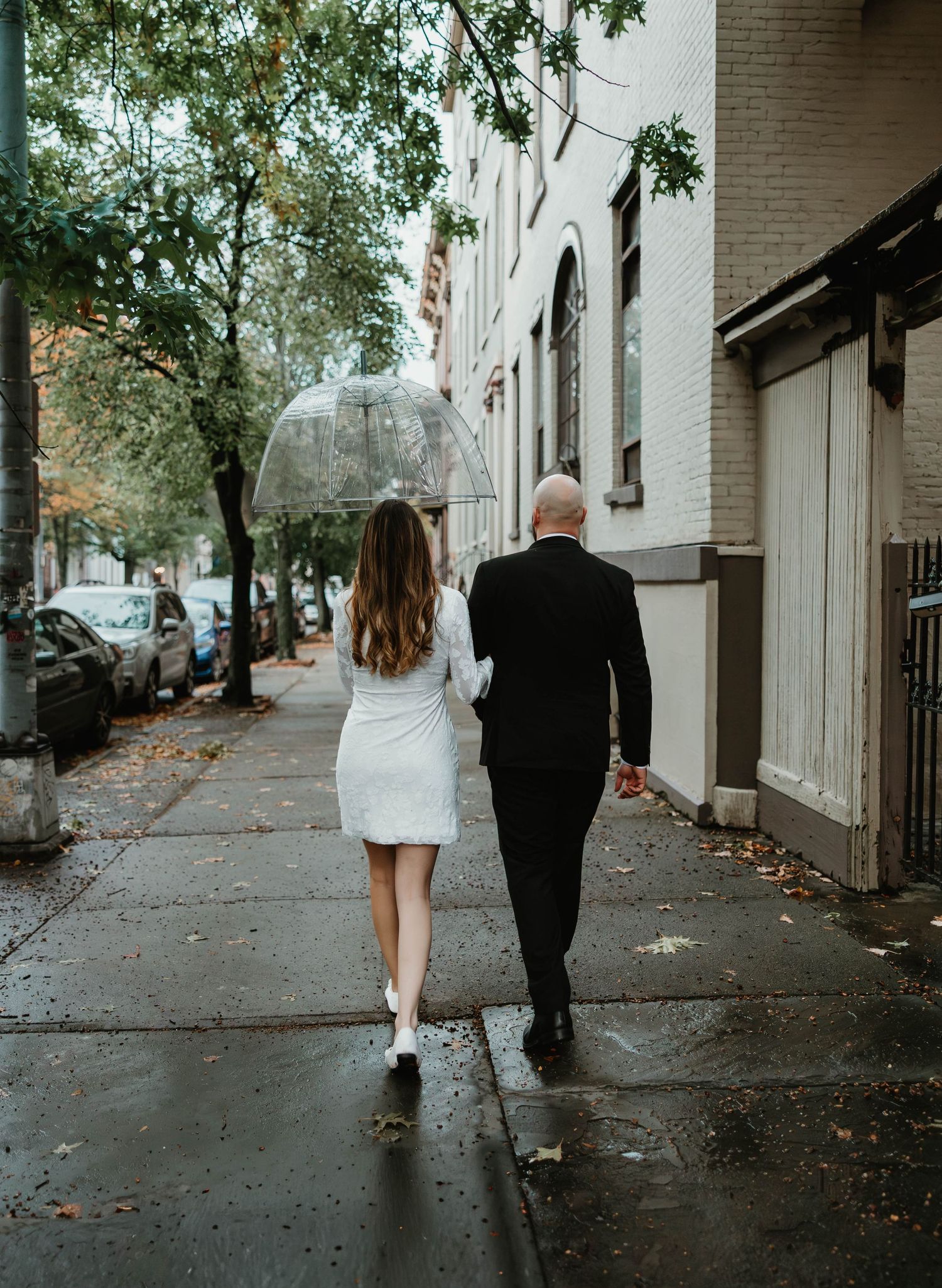 A couple walks together in the rain under a clear umbrella on a wet city sidewalk lined with trees.