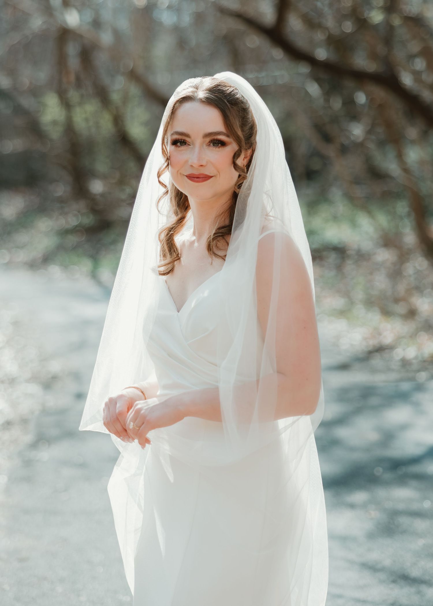 Bride in white wedding gown and veil poses outdoors among trees in soft natural light.