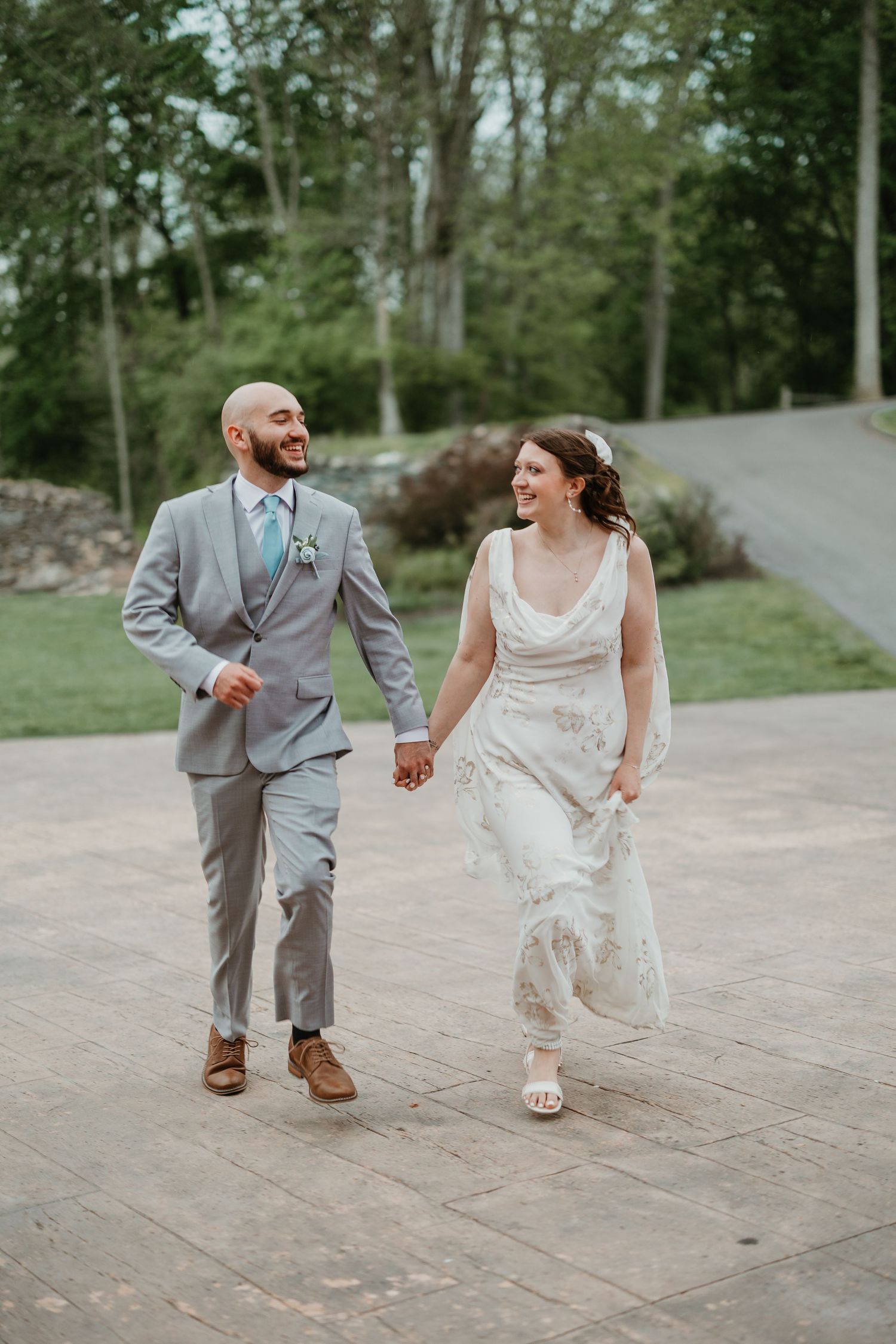 A smiling bride and groom hold hands while walking outdoors, she in a white gown and he in a gray suit with teal tie.