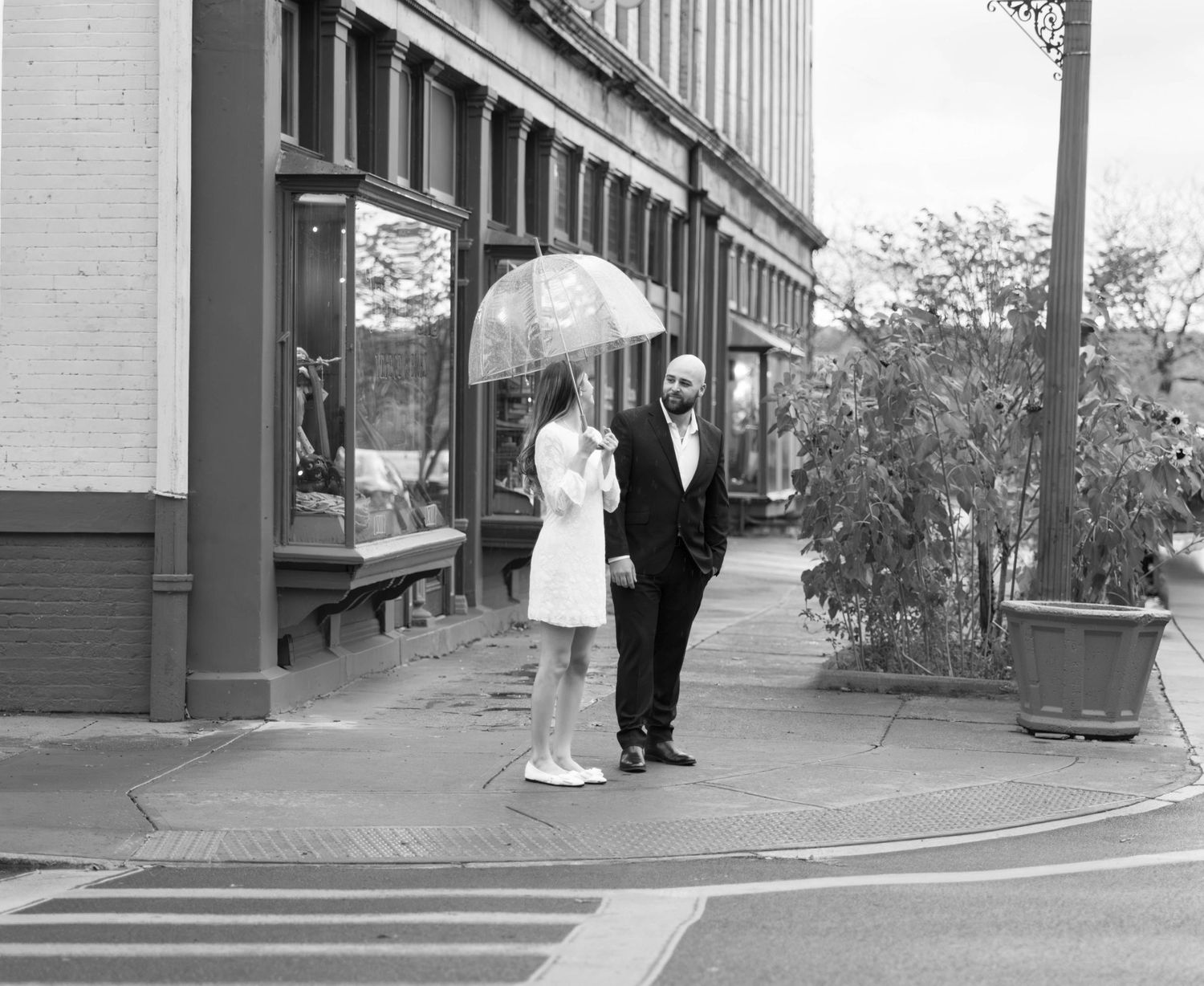 A black and white panoramic view shows couples walking along a sidewalk near storefronts in a downtown area.