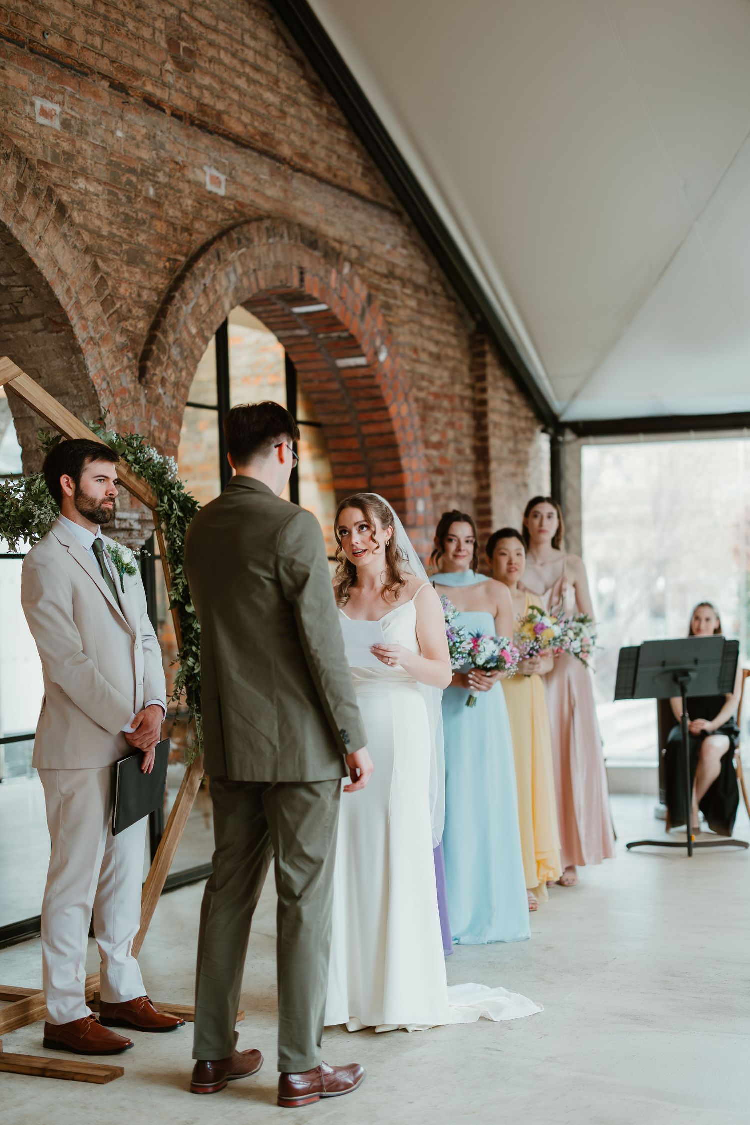 Bride and groom exchange vows at elegant indoor wedding ceremony with brick arches and colorful bridesmaids.