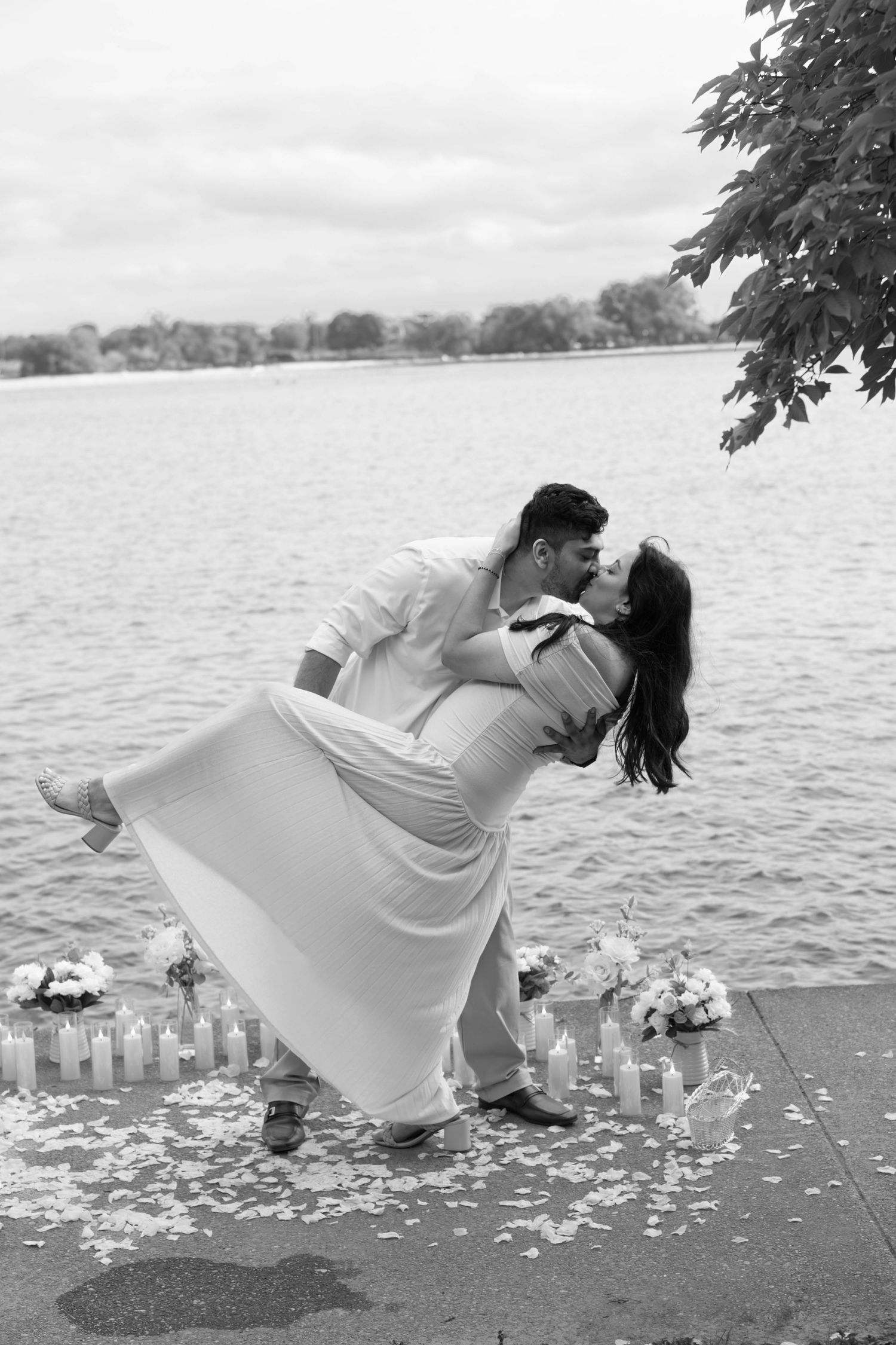 A romantic couple dips in an elegant dance pose by a lakeside with scattered flower petals on the ground.