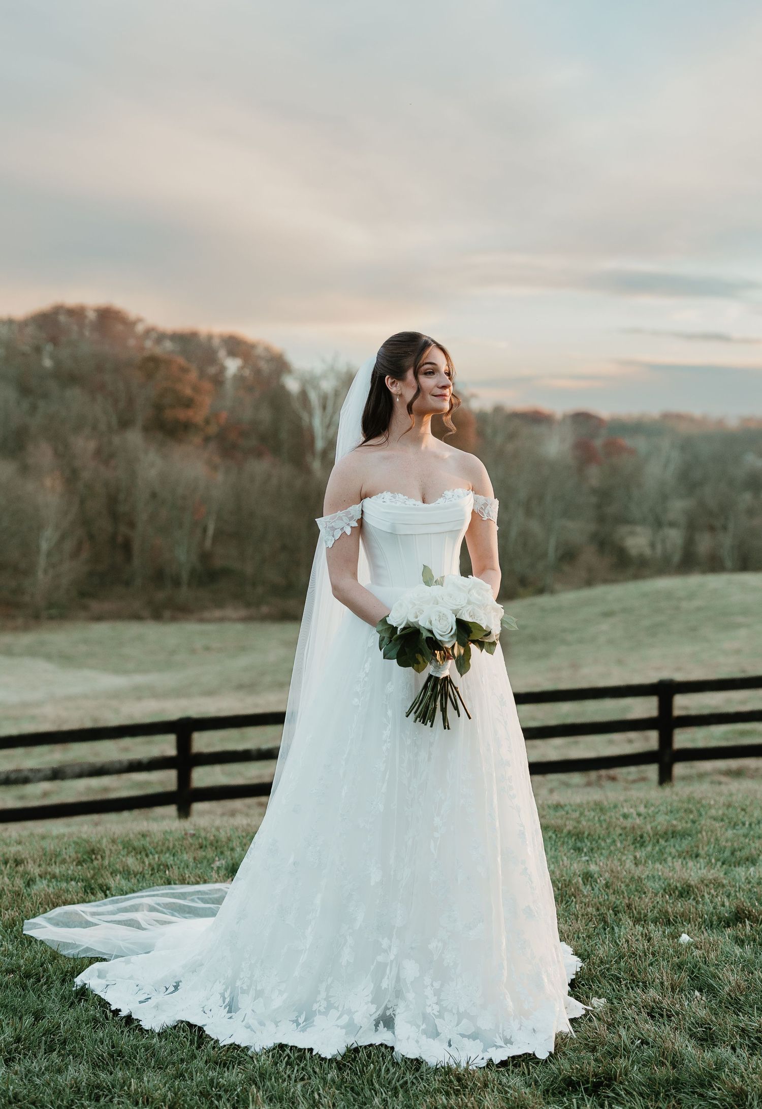 Bride in off-shoulder lace A-line gown with veil and bouquet, posing at golden hour on a green countryside estate.
