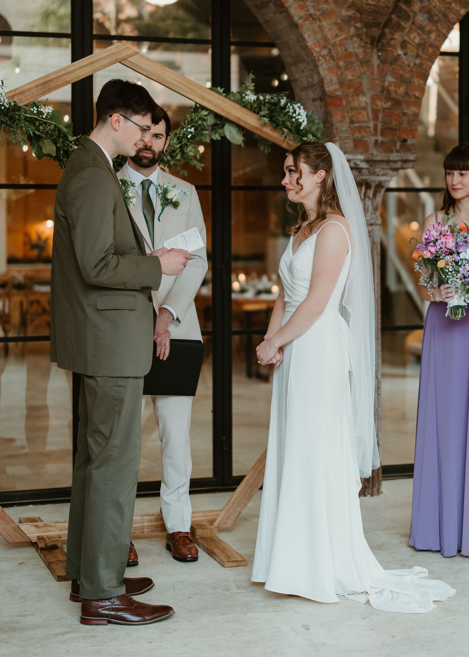 Bride and groom exchange vows at indoor wedding ceremony with wooden arch and greenery backdrop.