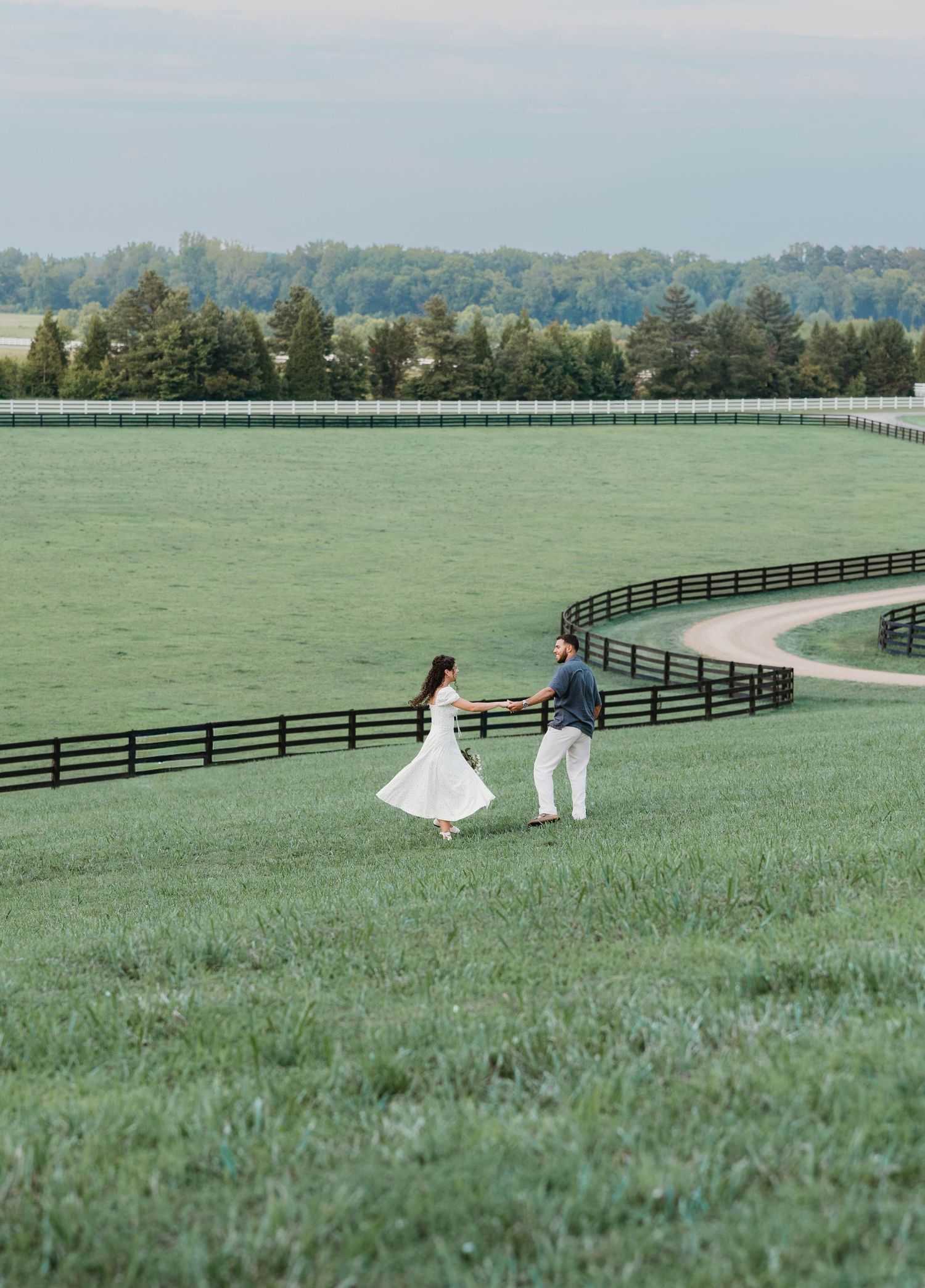A couple walks along a curved path through lush green pastures bordered by black fencing at a scenic horse farm.