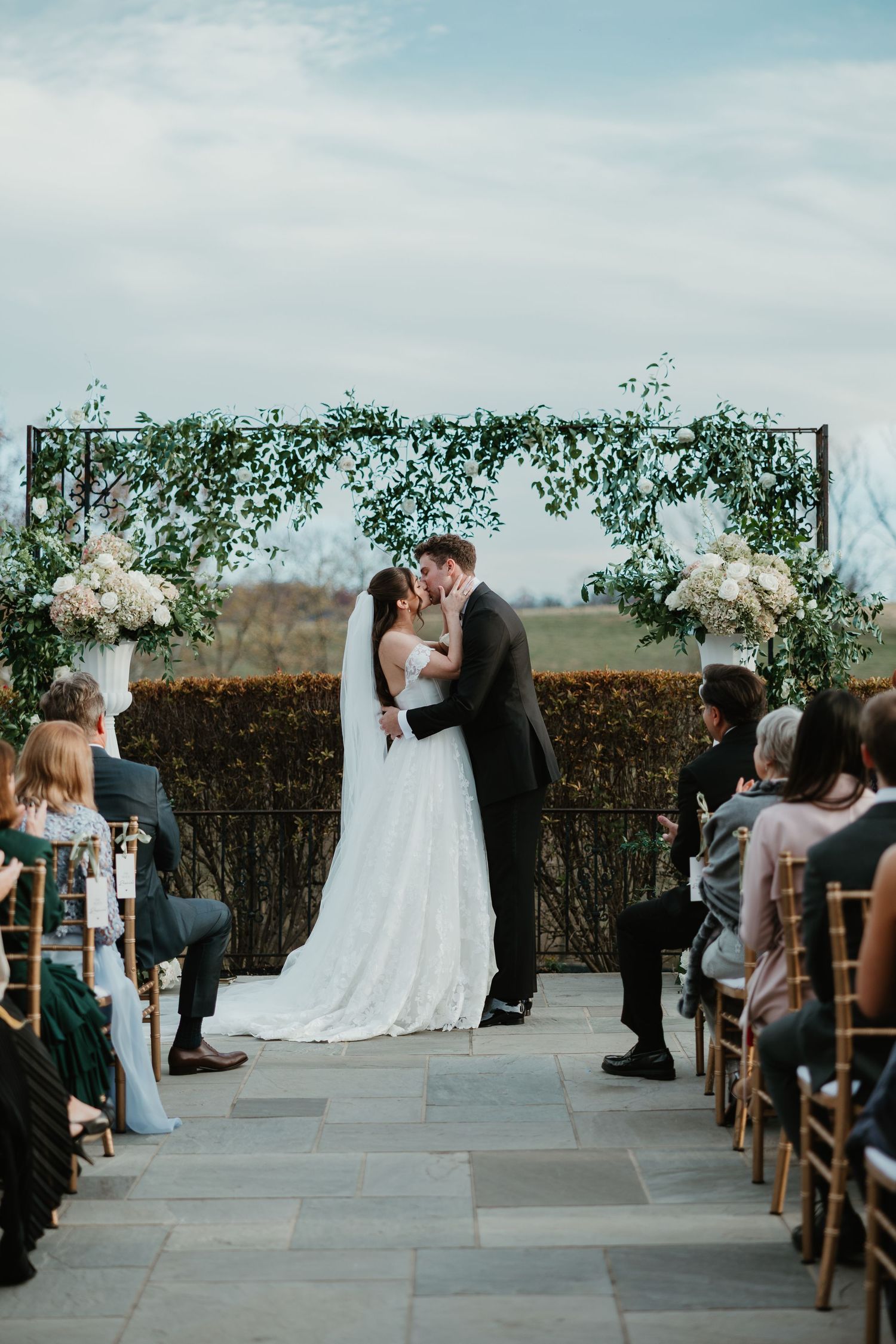 Bride and groom share first kiss at outdoor wedding ceremony beneath floral arch with white roses and greenery.