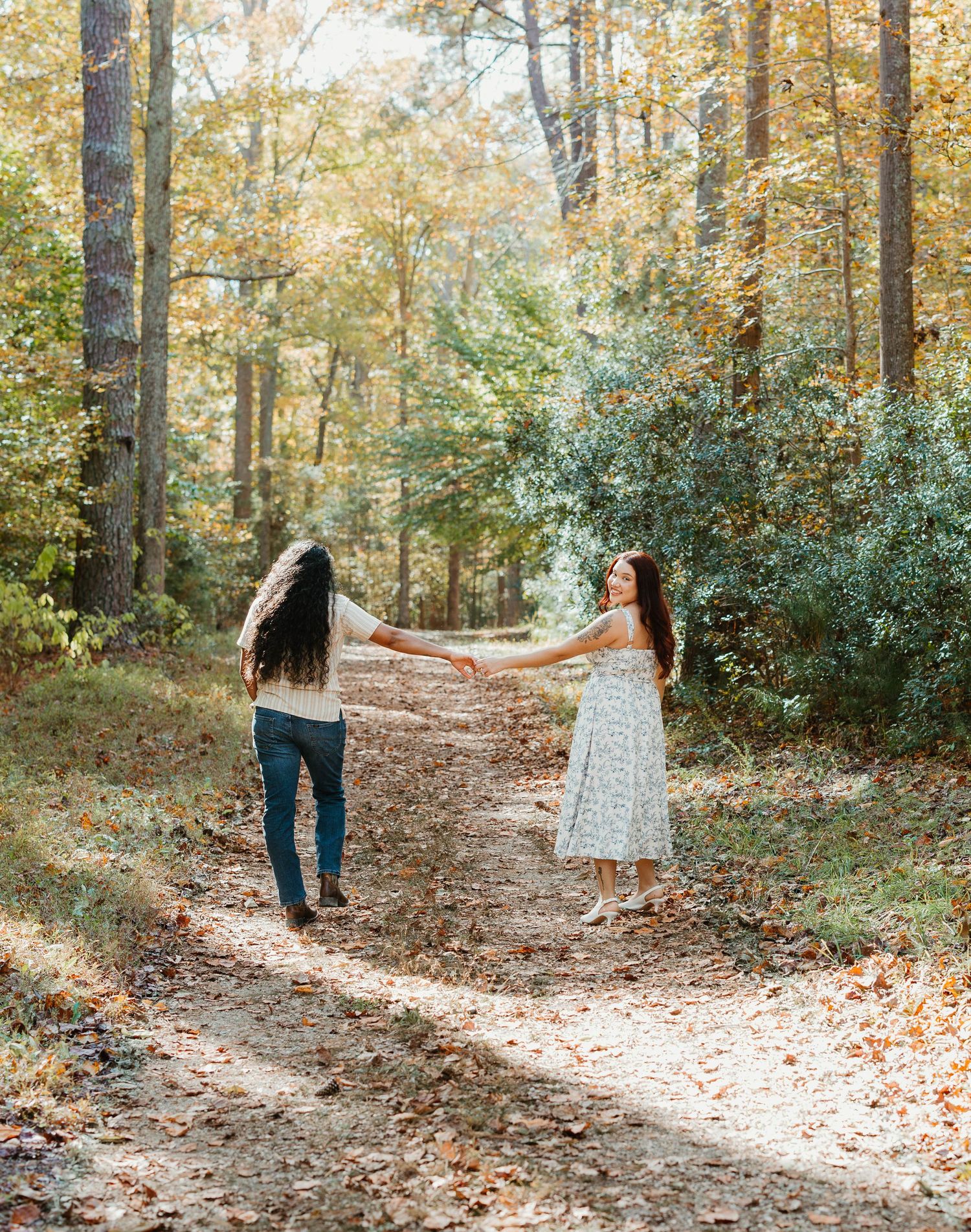 A couple holds hands while walking along a scenic autumn forest path covered in fallen leaves.