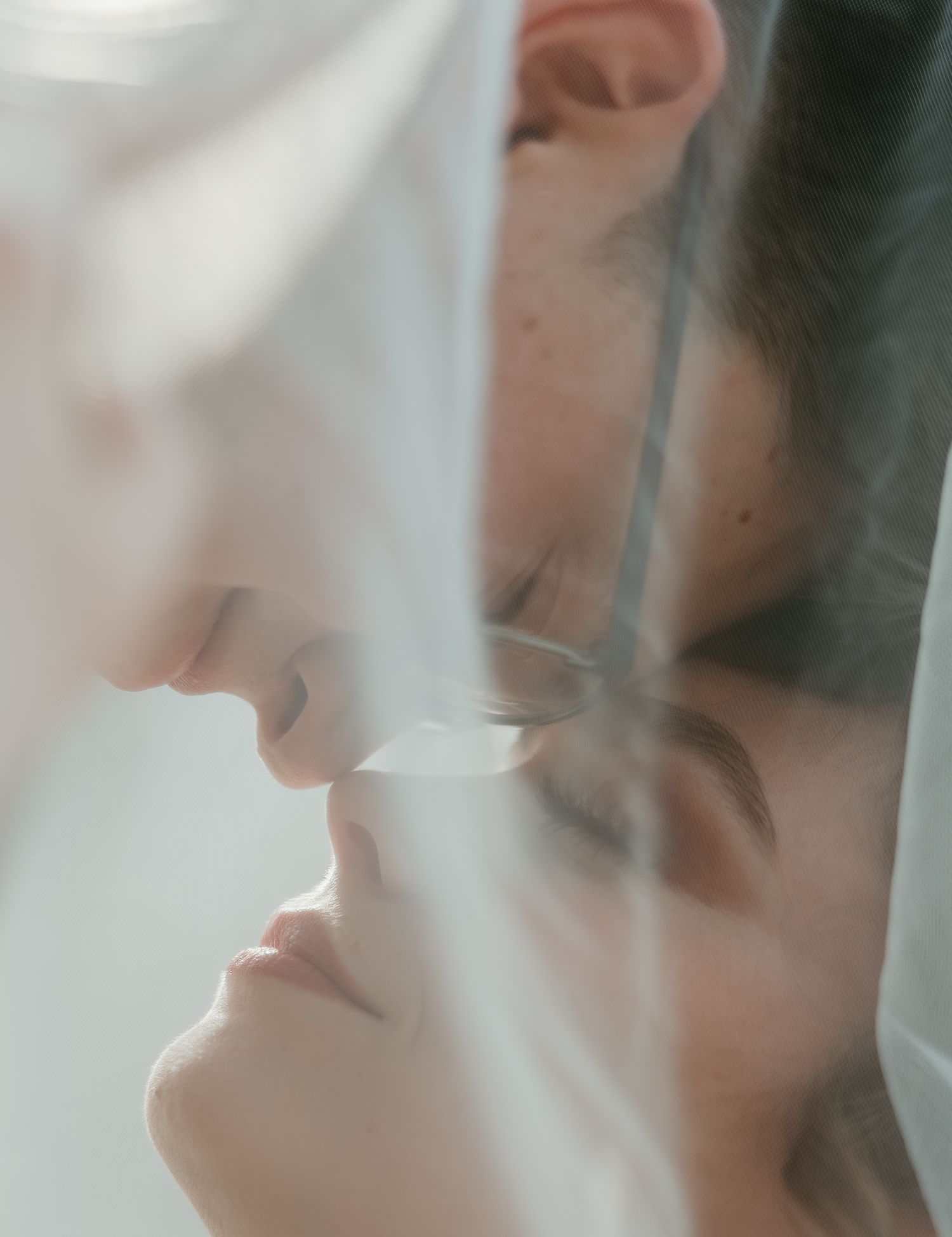 Two people sharing a tender almost-kiss behind a sheer white veil in a romantic and intimate close-up portrait.