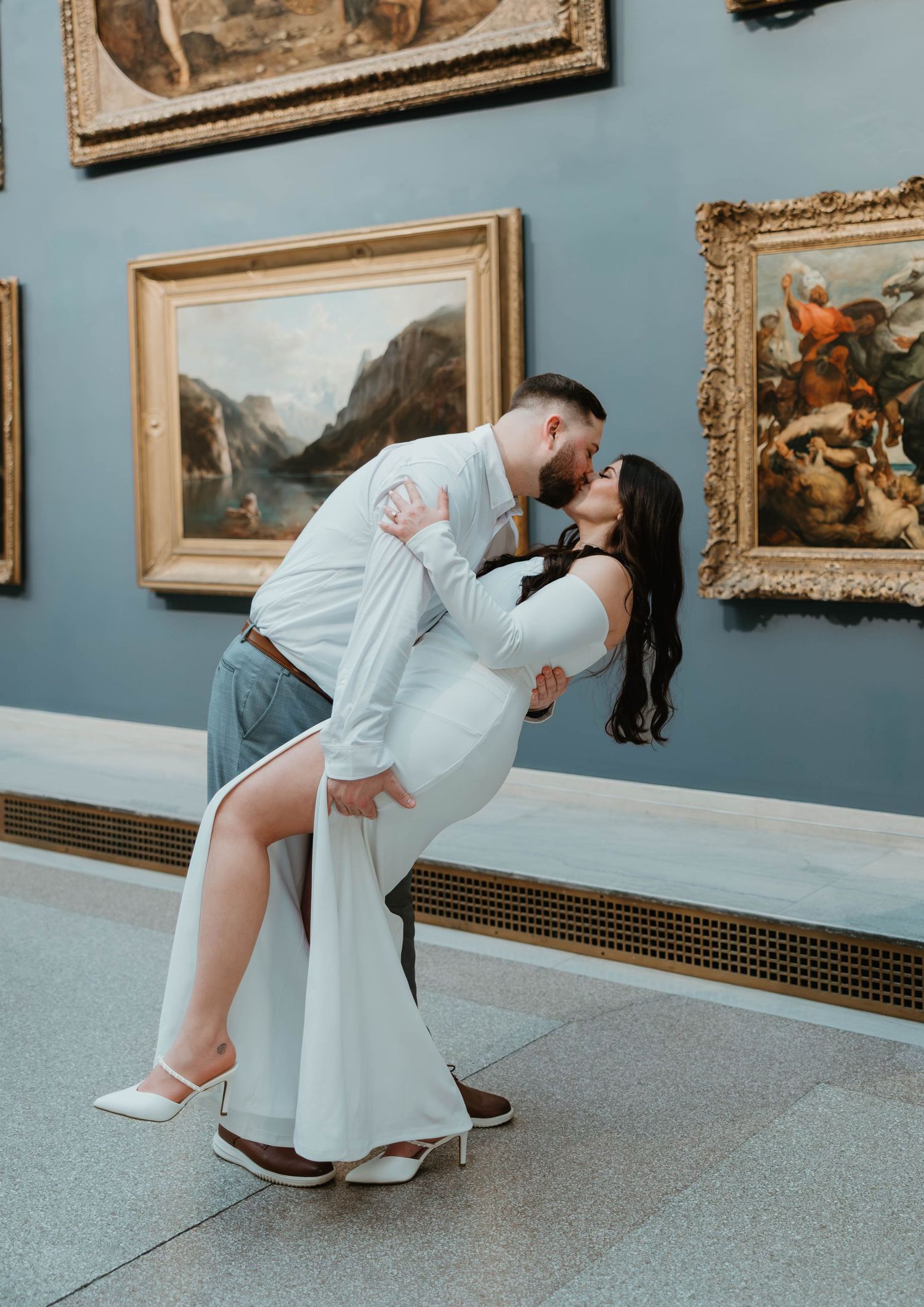 A person in a white dress crouches down to adjust their shoe in an art gallery with framed paintings on blue walls.