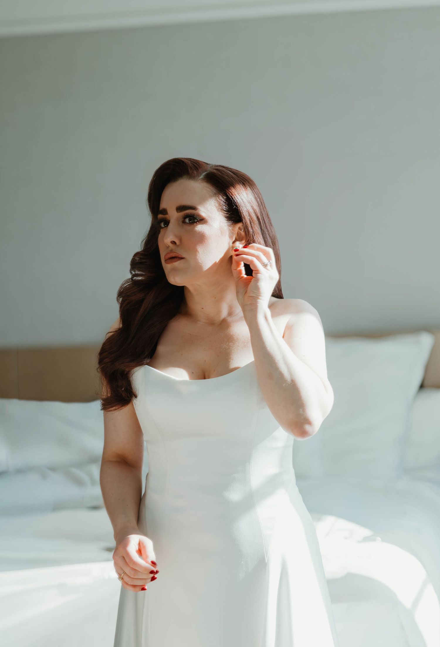 Bride in white strapless gown adjusting earring while standing in sunlit bedroom.