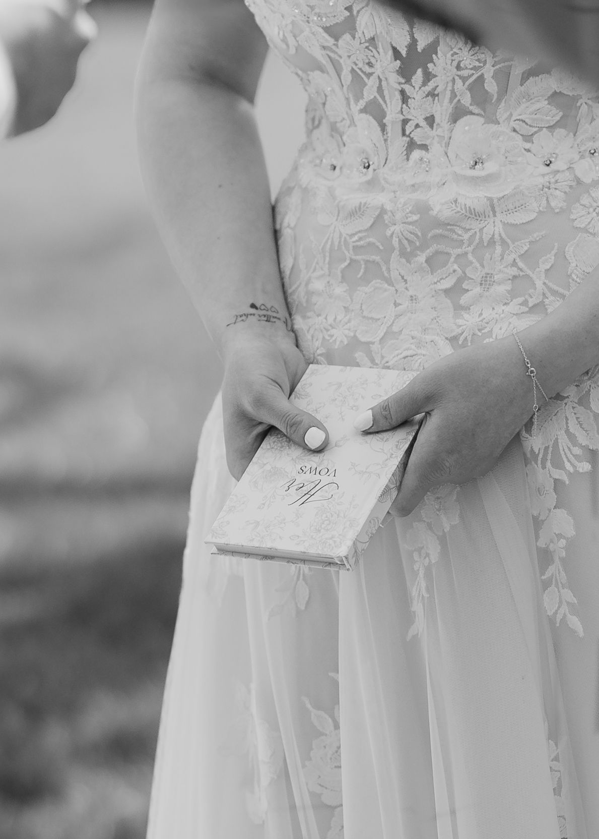 Bride in lace wedding gown holding elegant vow book during outdoor ceremony, black and white photo.