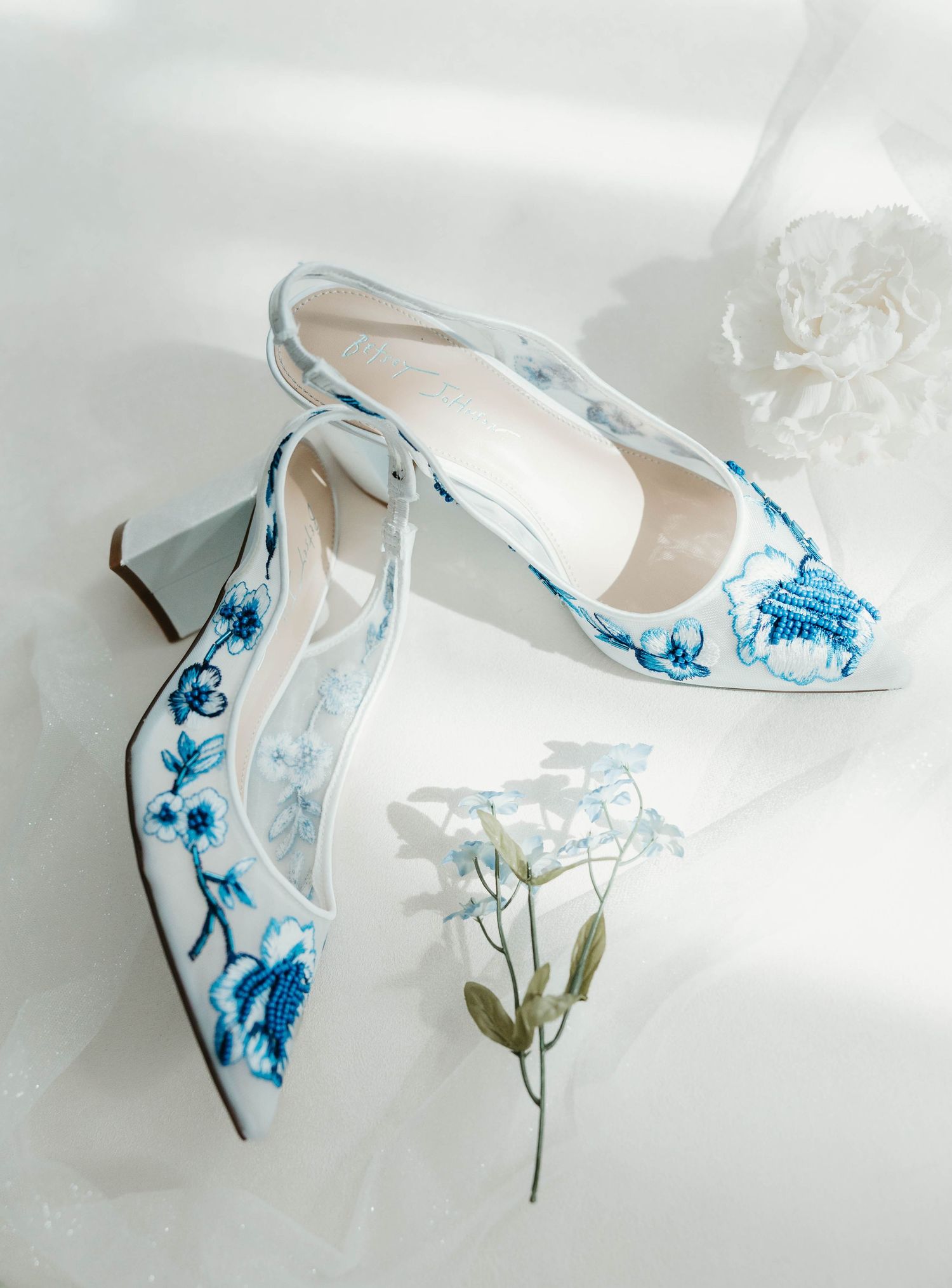 Elegant blue and white floral porcelain-inspired heels with delicate pattern details displayed with white flowers.