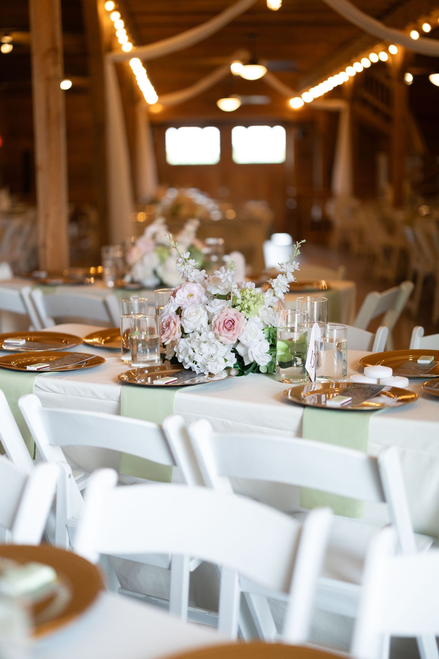 Elegant barn wedding reception with white chairs, gold charger plates, and blush floral centerpieces on linen-draped tables.