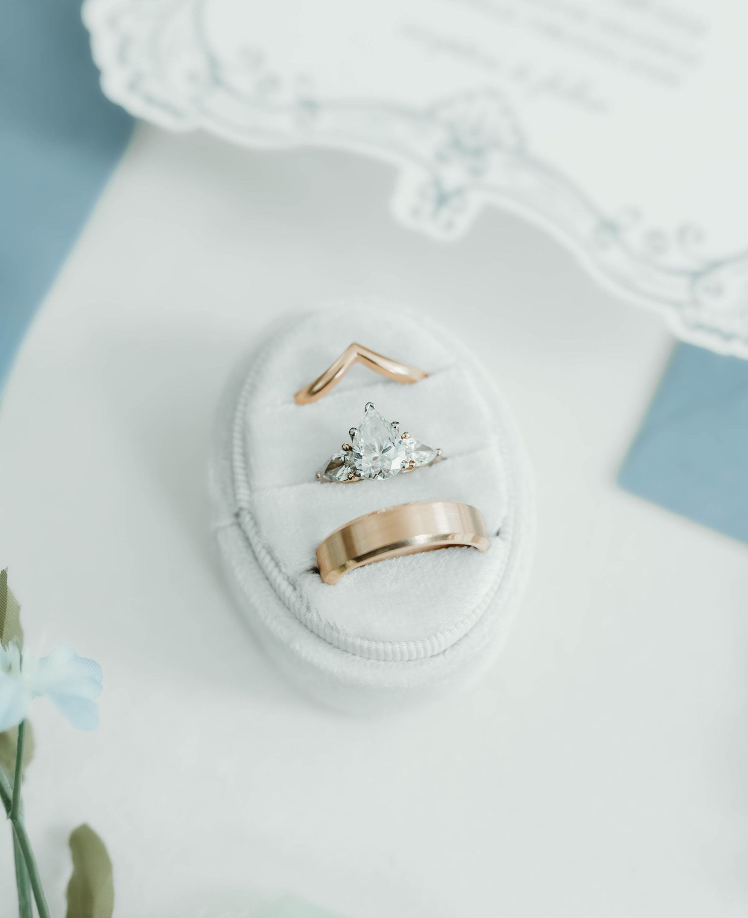 A delicate engagement ring with a diamond solitaire rests on a white textured surface next to floral elements.