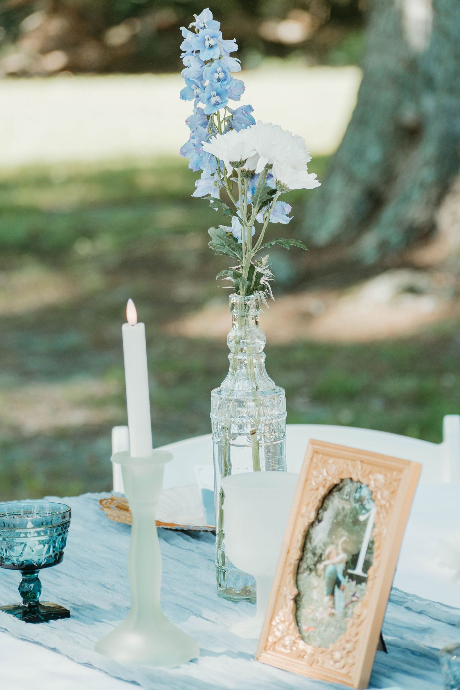 Vintage-style framed photo and delicate blue flowers in glass vases create an elegant table setting with white candles.