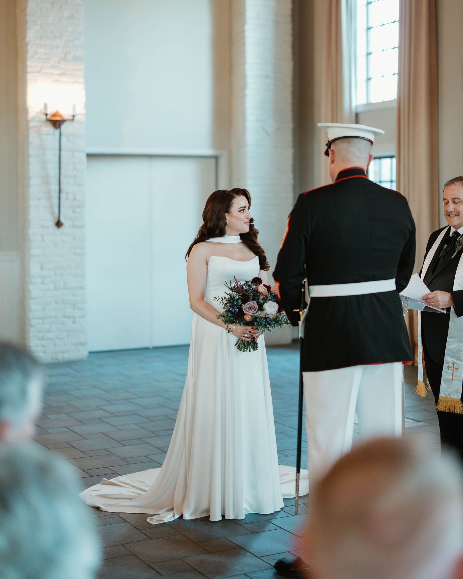 Bride in white off-shoulder gown holds bouquet facing groom in Marine dress uniform during wedding ceremony.