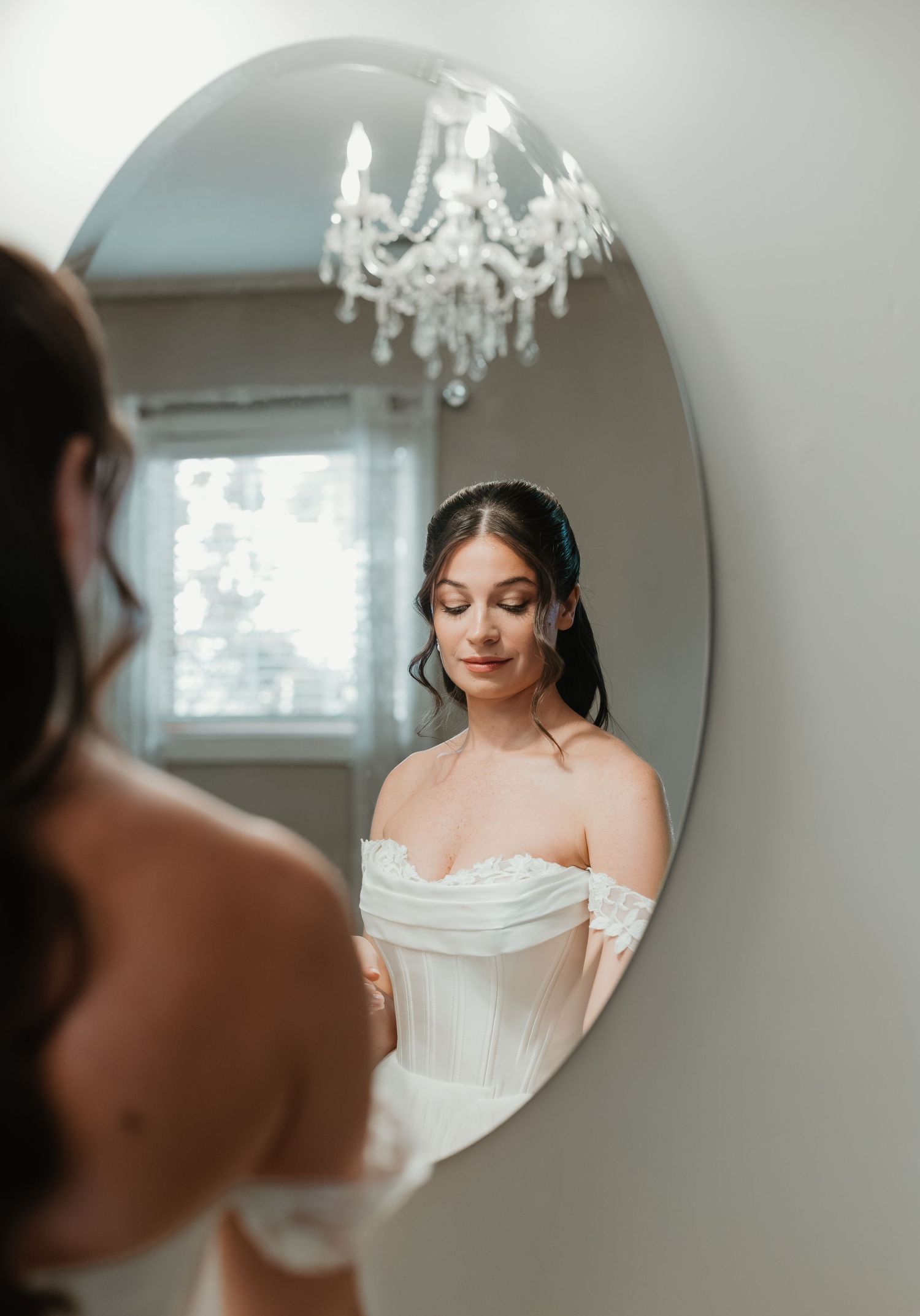 Bride in off-shoulder white wedding gown admires her reflection in an oval mirror beneath an elegant crystal chandelier.