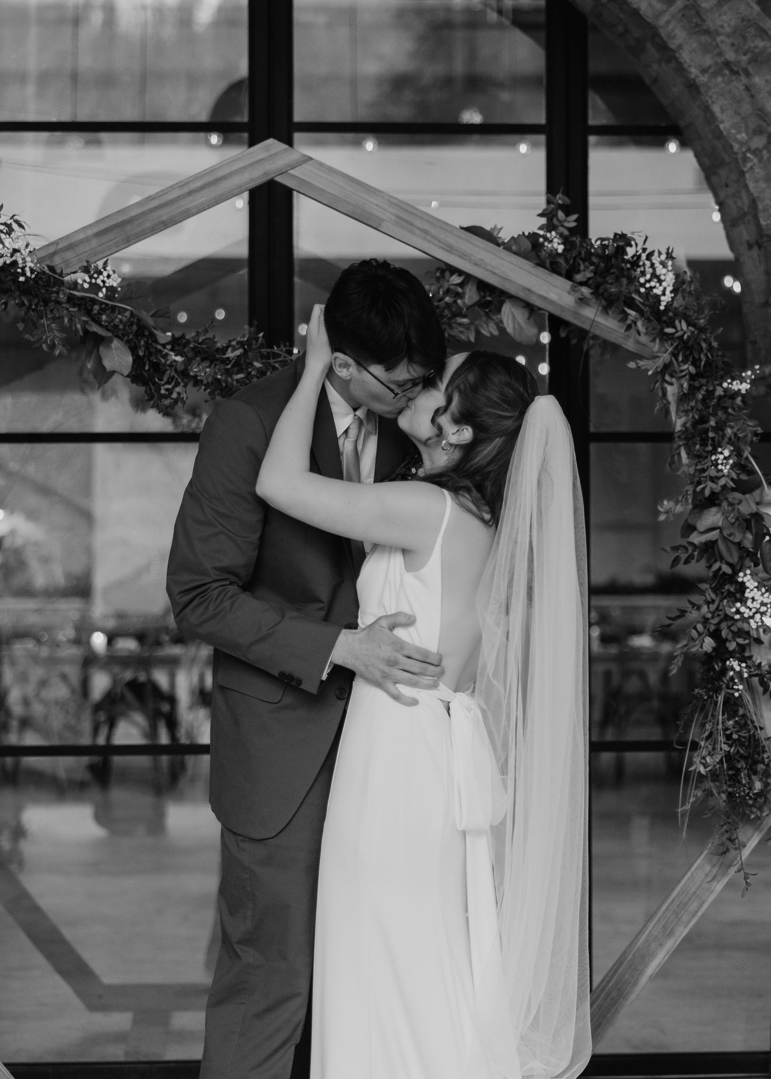 Black and white photo of a bride and groom sharing their first kiss under a wooden arch with greenery.