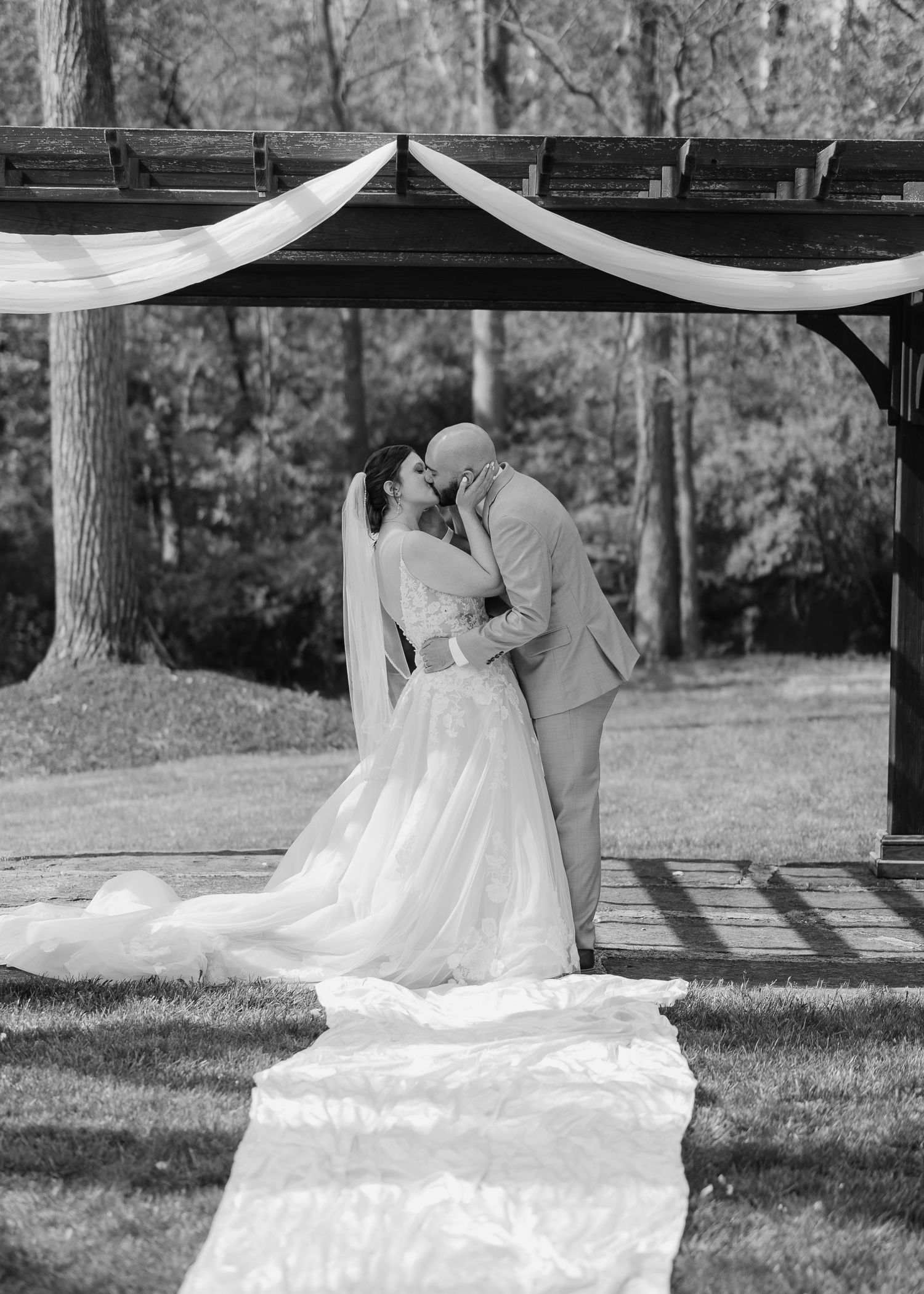 Black and white photo of a bride and groom sharing their first kiss under a wooden pergola draped with white fabric outdoors.