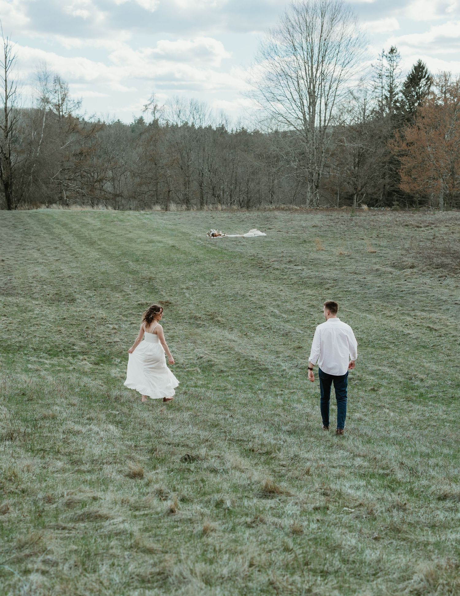 A couple walks hand-in-hand through a lush green meadow surrounded by bare trees during an outdoor engagement session.