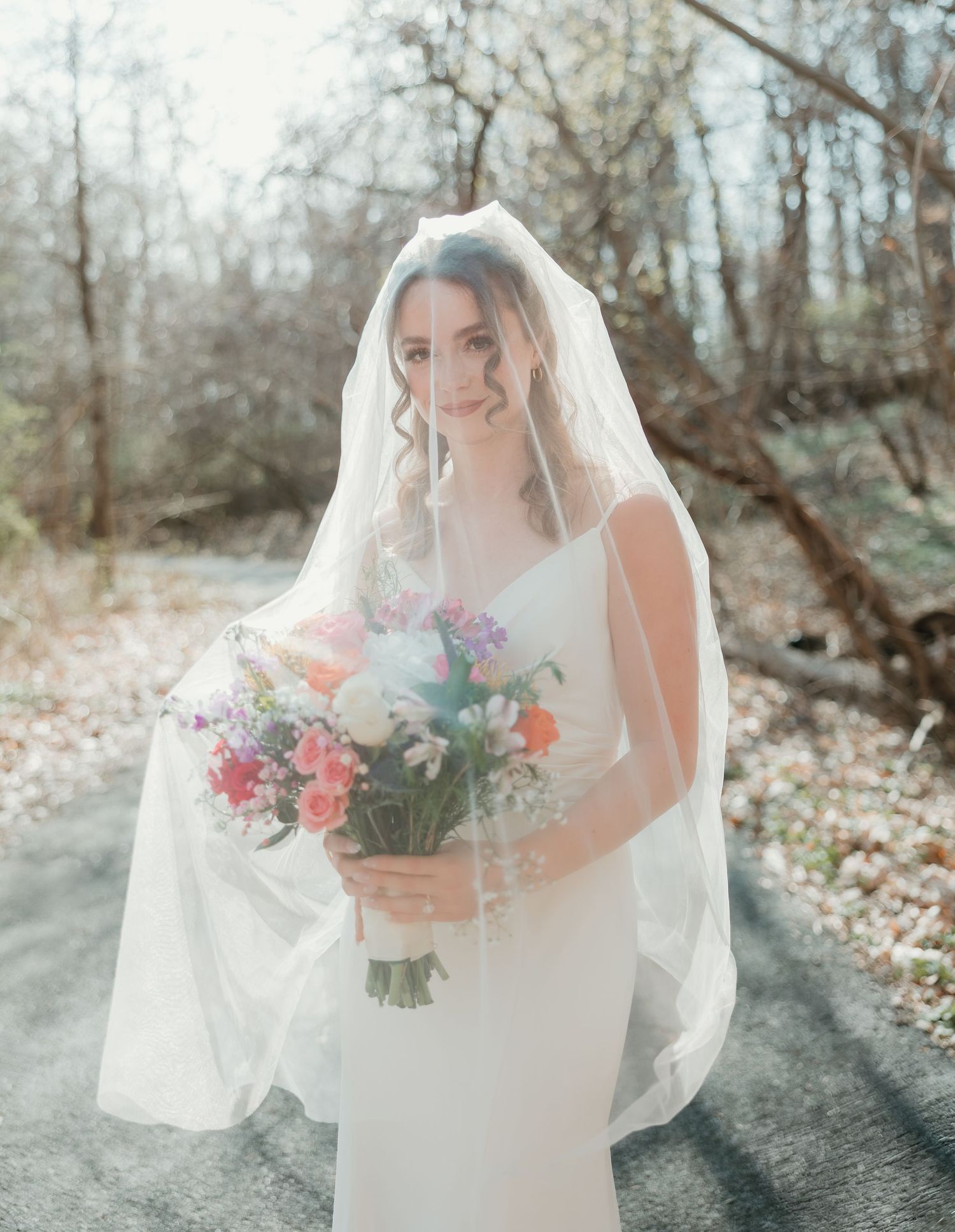 Bride in white gown and veil holding colorful bouquet of flowers outdoors in a wooded setting.