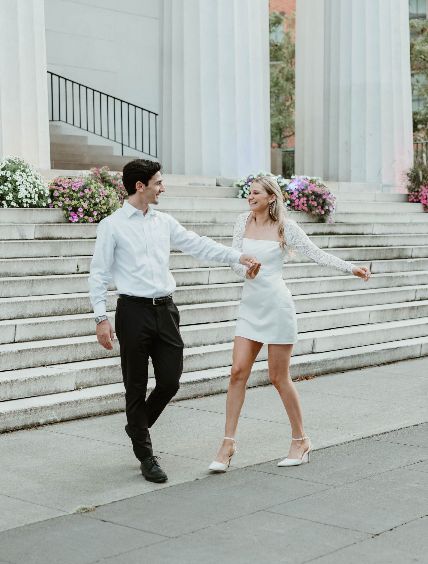 A couple in wedding attire dances playfully on marble steps adorned with pink floral arrangements.