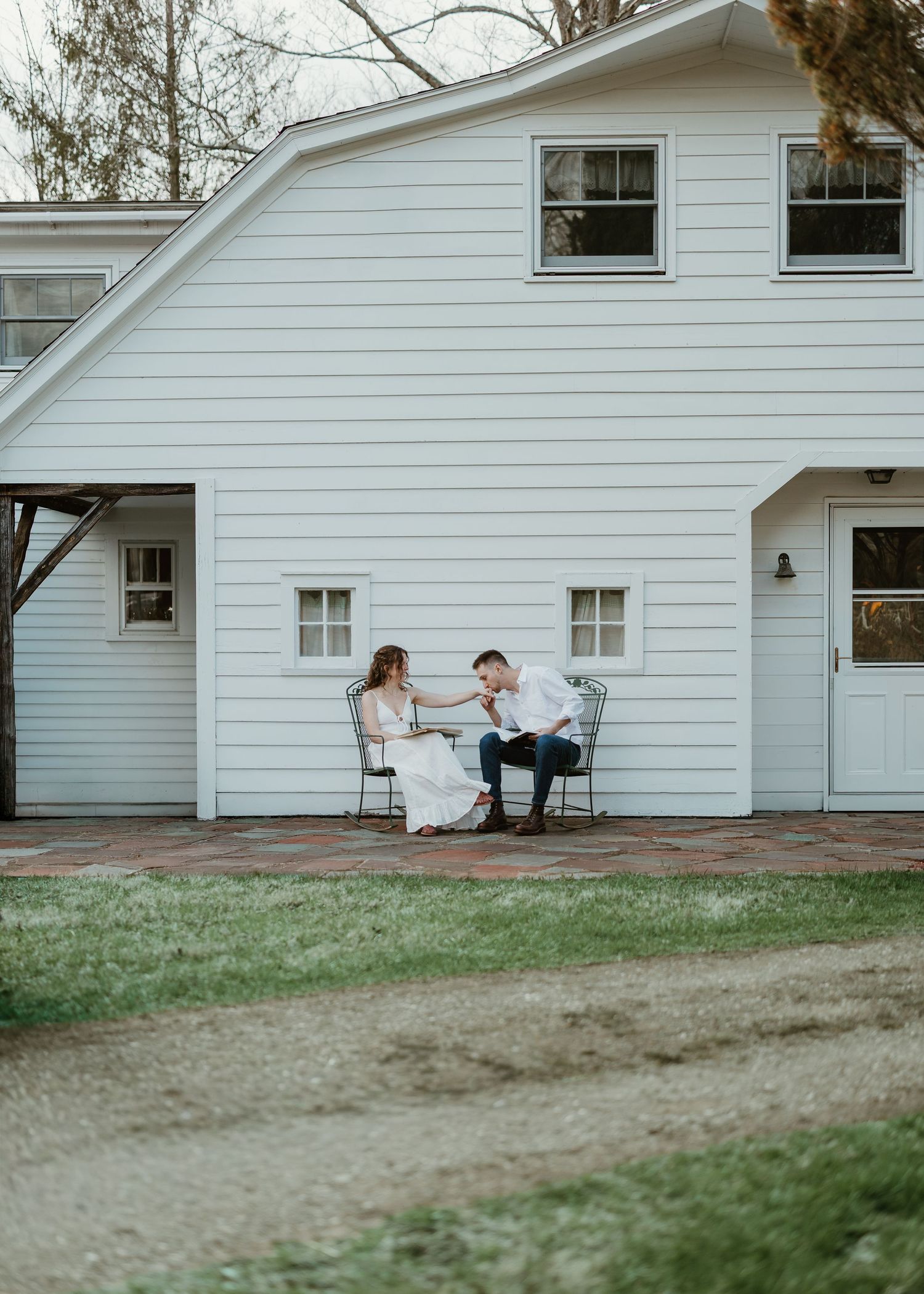 A couple in wedding attire sits on chairs outside a white farmhouse barn, sharing an intimate moment on a brick patio.