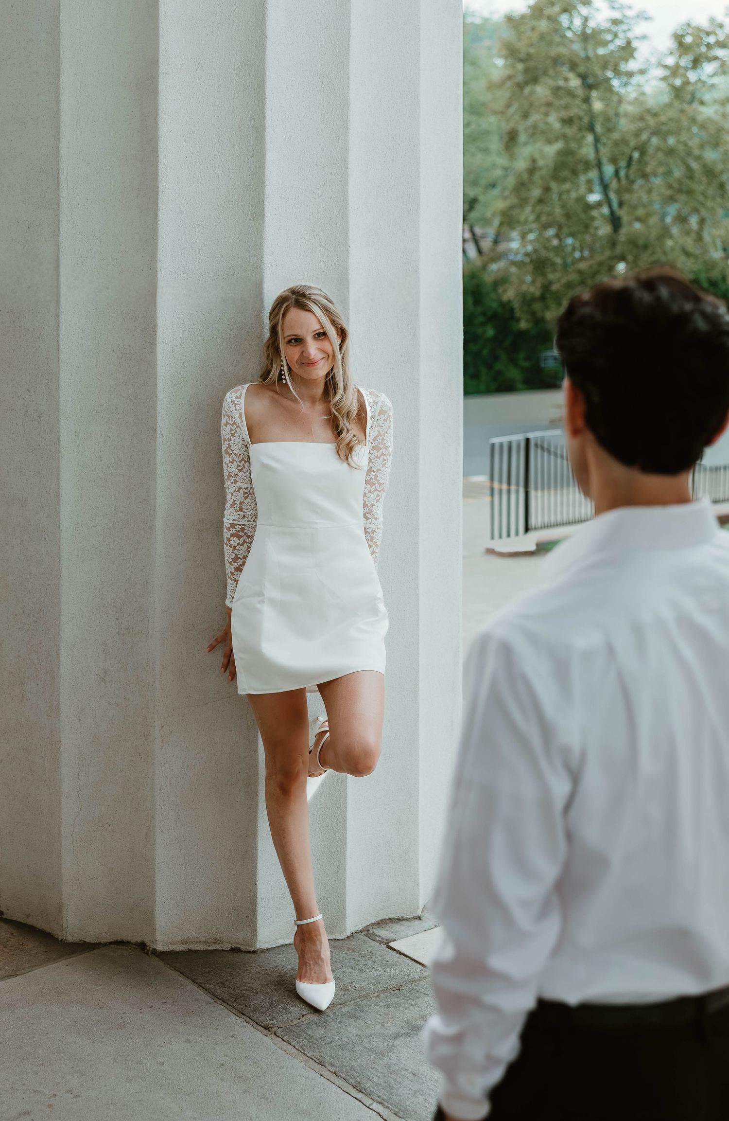 A romantic scene between a couple in formal attire playing hide and seek around white columns outdoors.