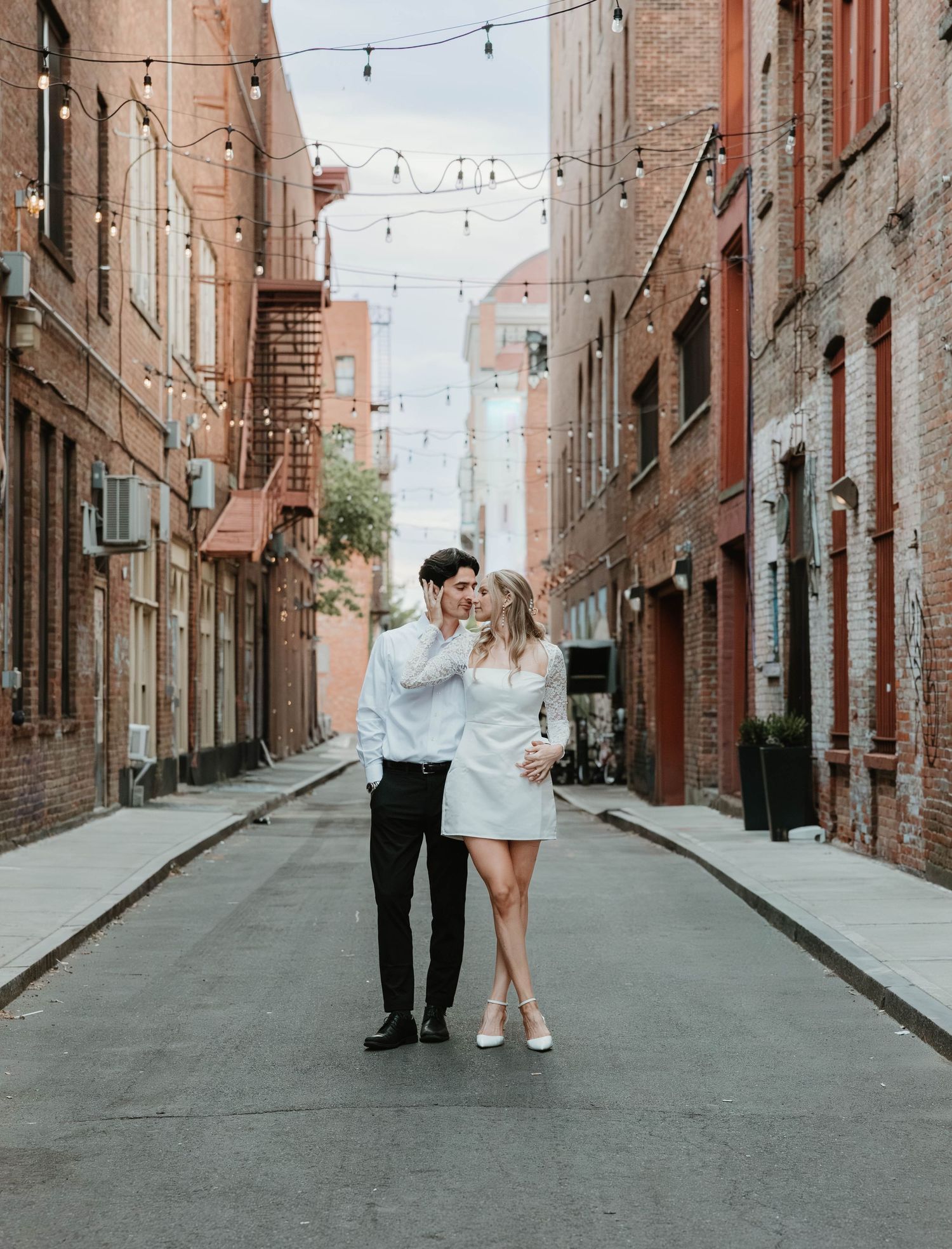 A romantic couple stands in the middle of a rustic brick alley with string lights hanging overhead during a sunset session.