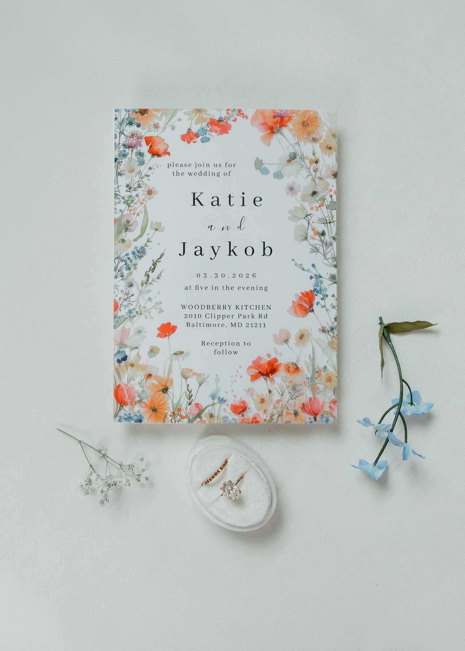 Wildflower wedding invitation for Katie and Jaykob with floral border, displayed with delicate blooms and pearl earrings.