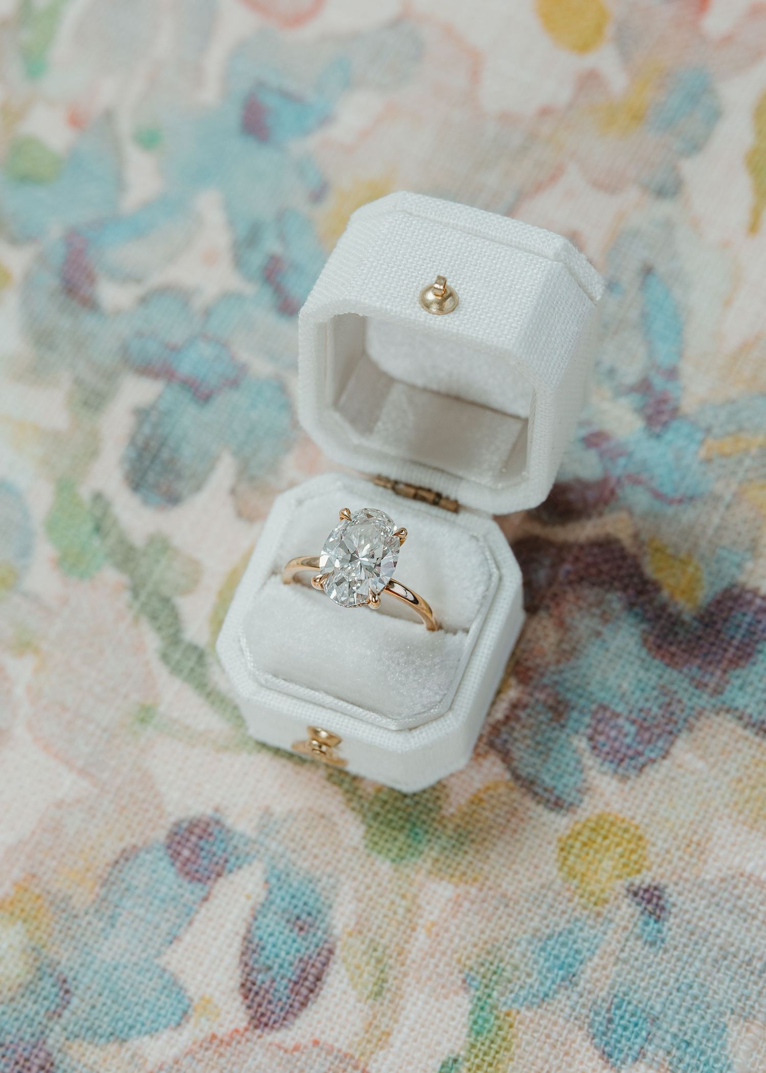 Oval diamond solitaire engagement ring in gold setting displayed in an open white velvet ring box on floral background.