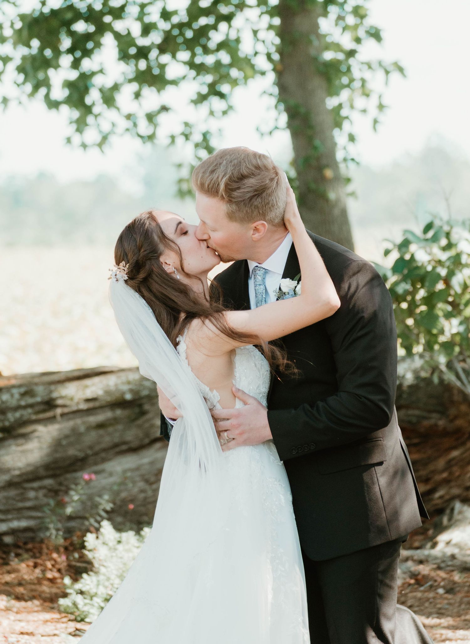 A romantic wedding couple shares tender kisses in a sunlit outdoor setting with trees in the background.