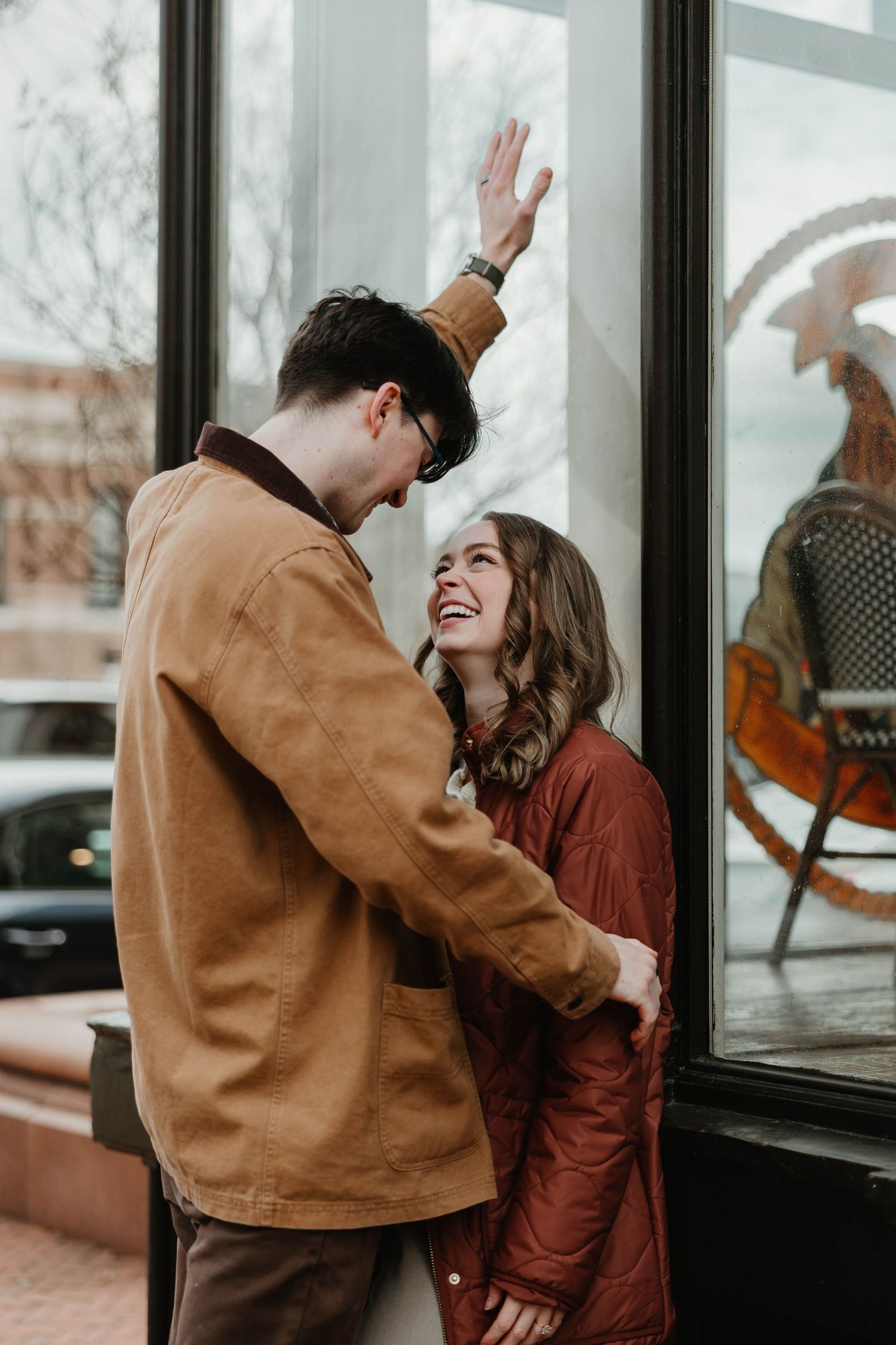 A couple laughs together in a candid moment by large windows, wearing brown and rust-colored jackets.