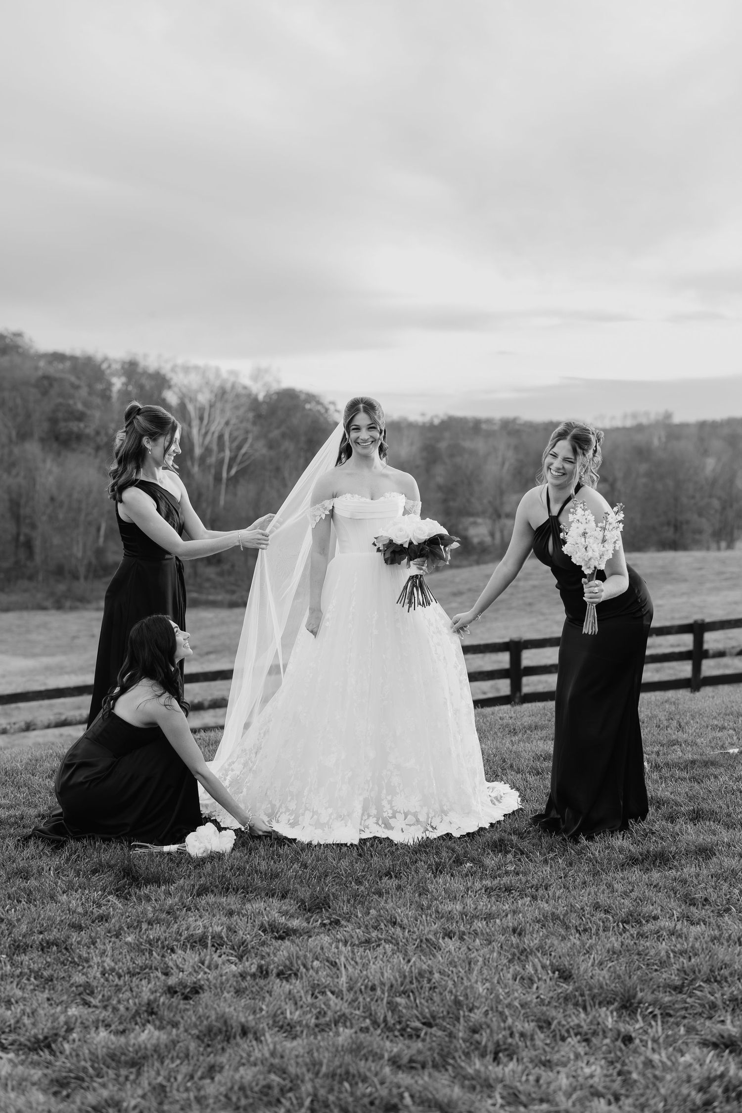 Bride in white gown and veil holds bouquet as bridesmaids in black dresses arrange her train outdoors.