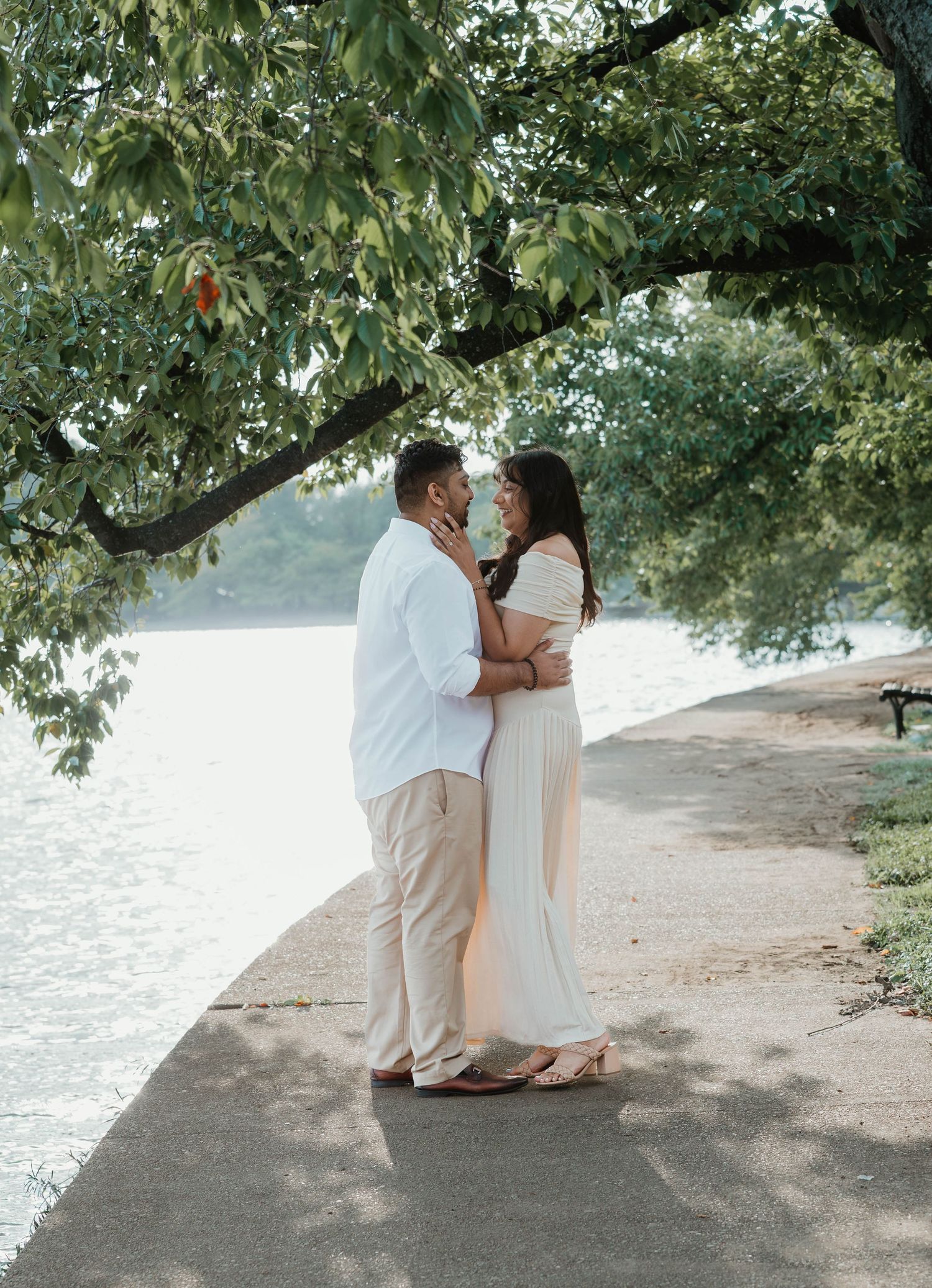A romantic couple embraces on a waterfront path under a leafy tree on a sunny summer afternoon.