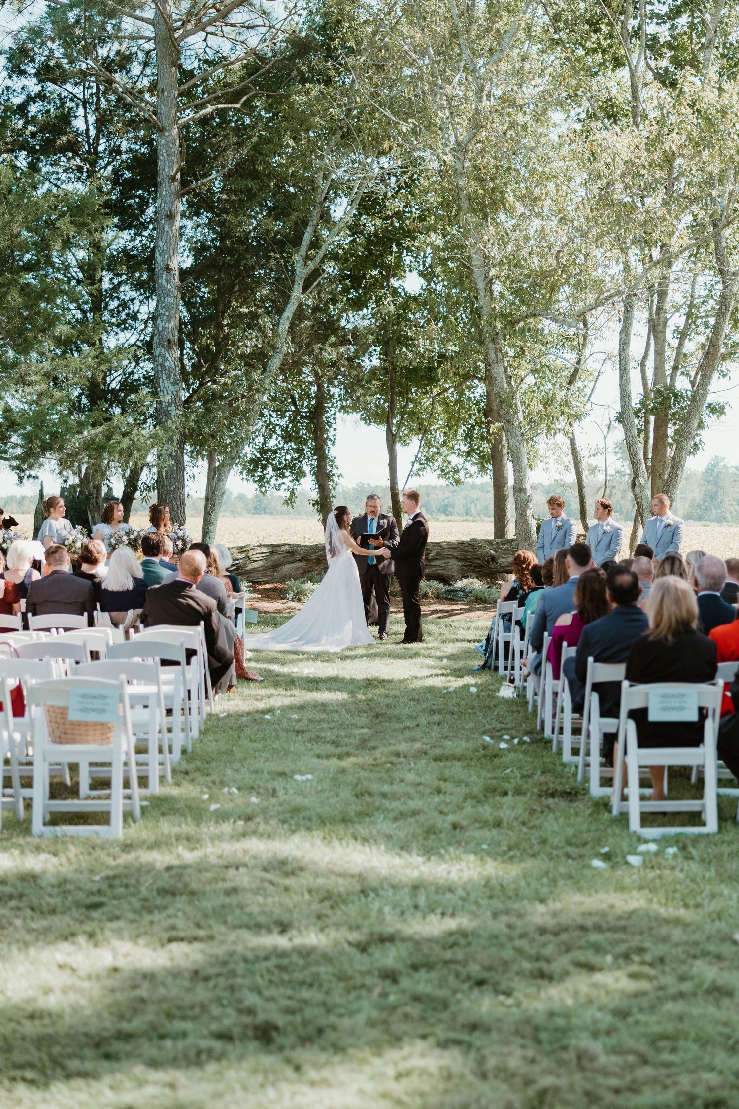 Outdoor wedding ceremony under tall trees with white chairs on grass and guests watching couple exchange vows.