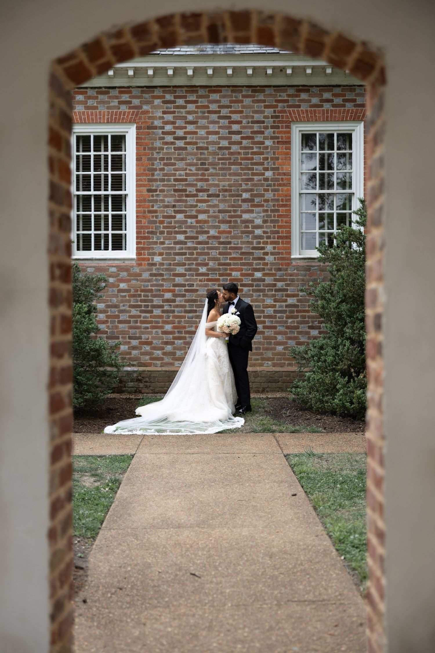 Bride and groom kiss outside a classic brick building, framed by an archway, with the bride's long veil trailing behind.