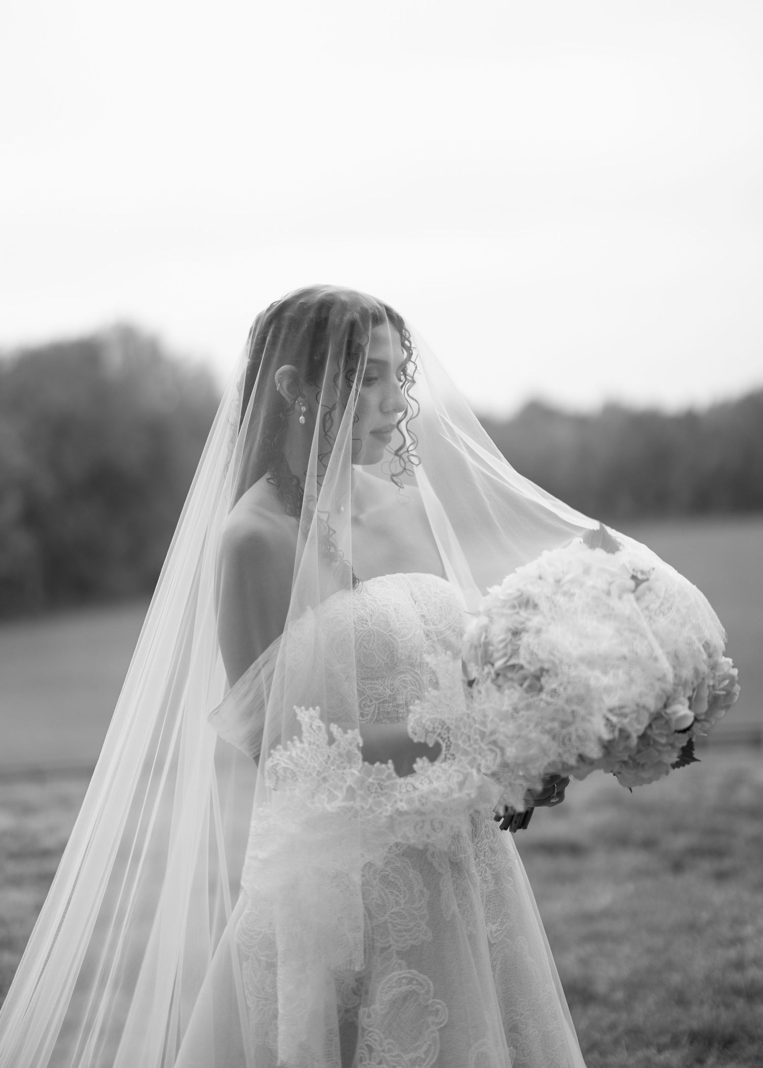 Black and white bridal portrait of a bride in a lace gown and flowing veil holding a white floral bouquet outdoors.